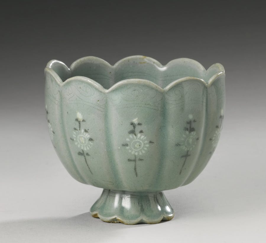 A Korean slip-inlaid celadon stoneware cup, Goryeo Dynasty, 13th century. Ceramic with a foliate rim and ten lobes, supported on a flared stem foot, each lobe inlaid with white and black slip with a chrysanthemum spray between incised foliate scrolls and waves, the interior with an incised floral medallion. Photo © Sotheby’s