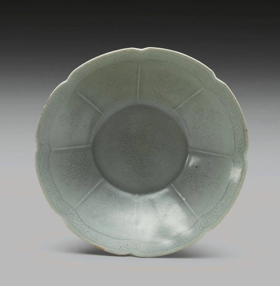 A Korean celadon foliate rim dish, Goryeo Dynasty, 12th century.  Ceramic with straight flaring sides rising to the notched rim, the interior divided into eight panels, each incised with a stylized lotus spray, beneath a foliate scroll at the rim, the well incised with a larger lotus spray, the incised sprays repeated on the exterior and base, all covered with a gray-green glaze, the base with three spur marks. Photo © Sotheby’s 