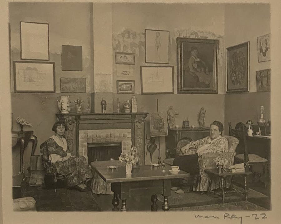 A photo of the photographic print by Man Ray titled Alice B. Toklas and Gertrude Stein (1922) at the National Gallery of Art in 2022. A black and white gelatin silver print of the named literary figures in Stein's salon in 1922. Below the image are Man Ray's signature and the year written in pencil. Public domain image.