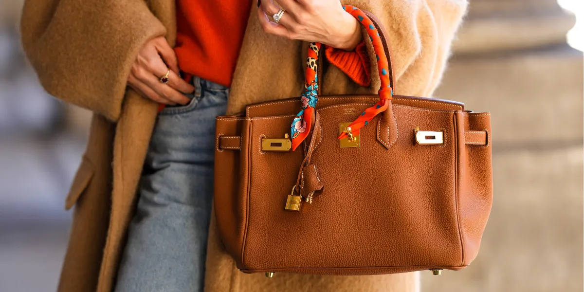 PARIS, FRANCE - FEBRUARY 09: Alexandra Pereira wears a neon orange turtleneck wool pullover, a beige oversized long winter coat, a brown shiny leather Birkin handbag from Hermes, diamonds ring, an orange / blue / yellow / white print pattern silk scarf from Hermes, blue faded denim large mom jeans pants, during a street style fashion photo session, on February 09, 2022 in Paris, France. Photo by Edward Berthelot/Getty Images (detail)