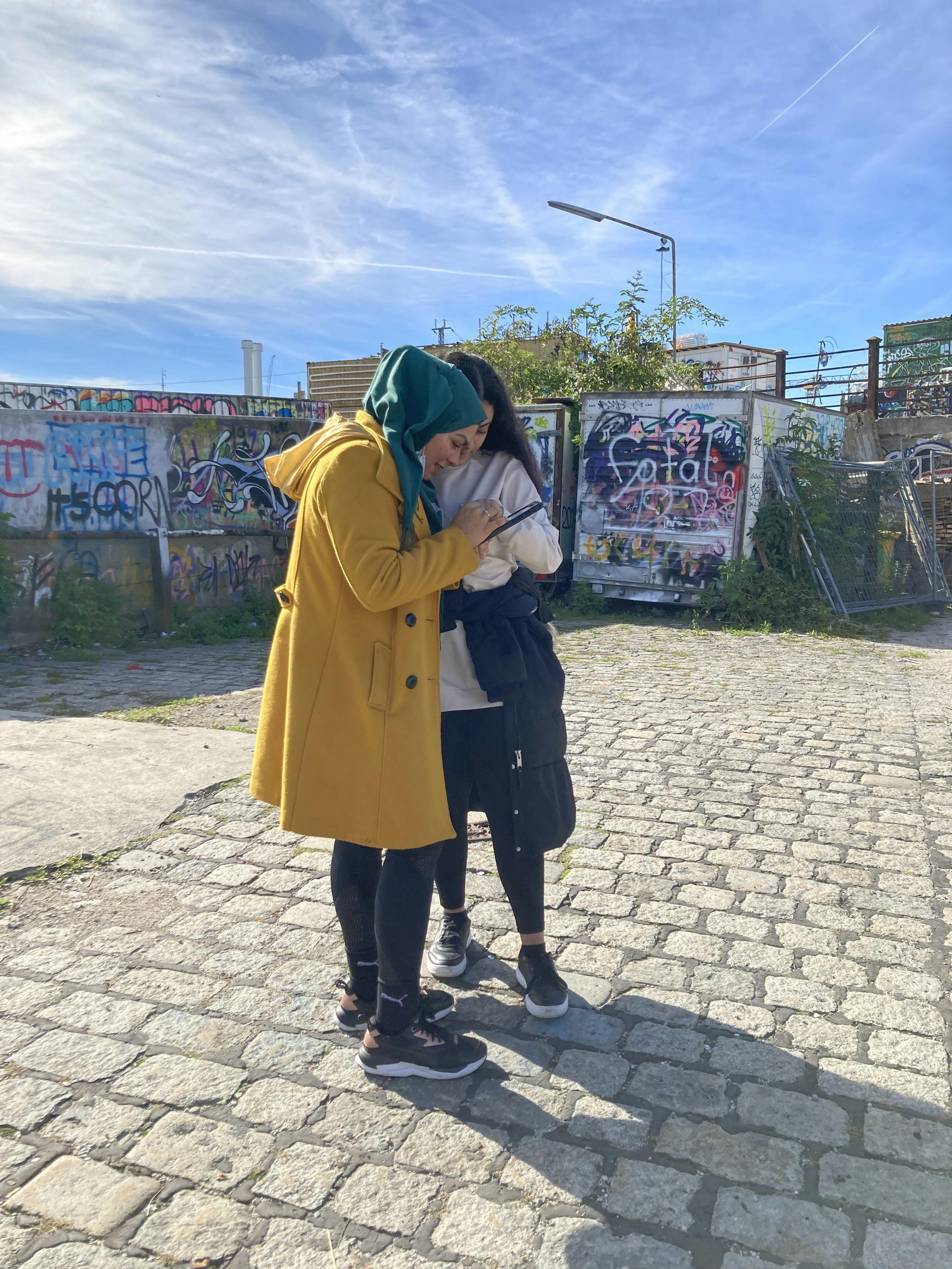 Two women play the augmented reality game Canvas City with a tablet. Walls with graffiti can be seen behind them.