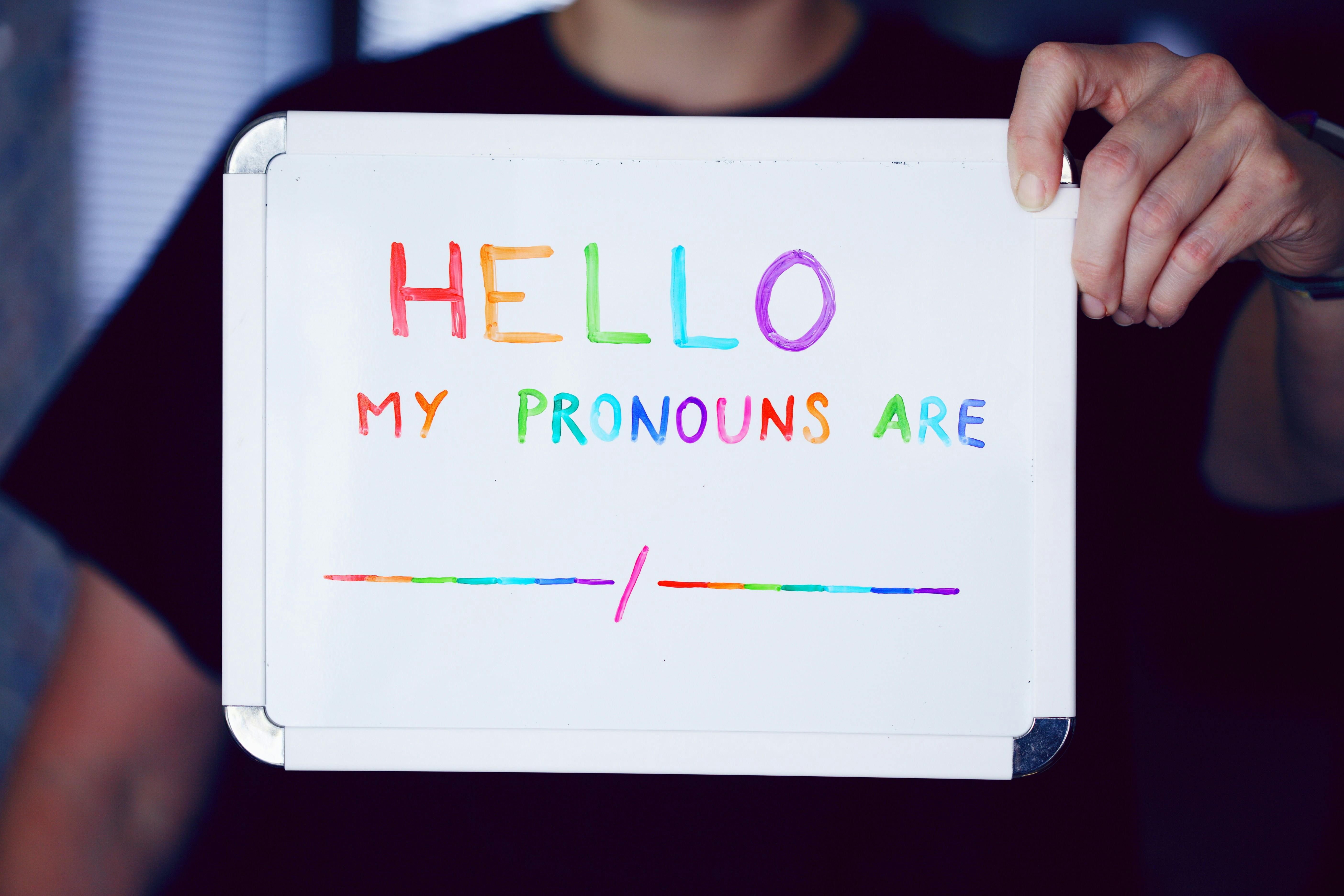 Eine Person hält eine Tafel in die Kamera auf der in regenbogenfarbener Schrift steht: Hello. My pronous are .../... 