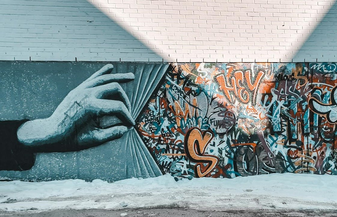 Auf dem Foto ist eine Wand mit einem Graffiti zu sehen. Die wand ist rechts komplett voll mit Graffiti. Links zieht eine riesige gemalte Hand einen gemalten Vorhang zur Seite, so dasss die ganzen Tags sichtbar werden. 