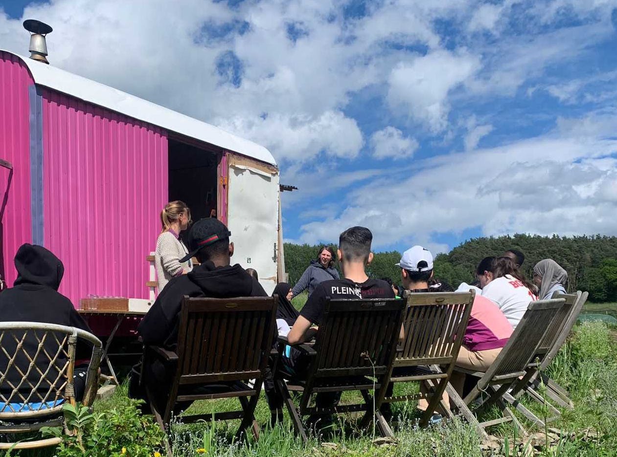 Auf dem Foto sieht man sitzend einige Teilnehmende des Seminars. Stehend im Hintergrund sind zwei der Referent:innen zu sehen. Den Bildhintergrund bildet der pinke Bauwagen der SoLaWi Stolze Gärtner und der blaue Himmer mit Schäfchenwolken.