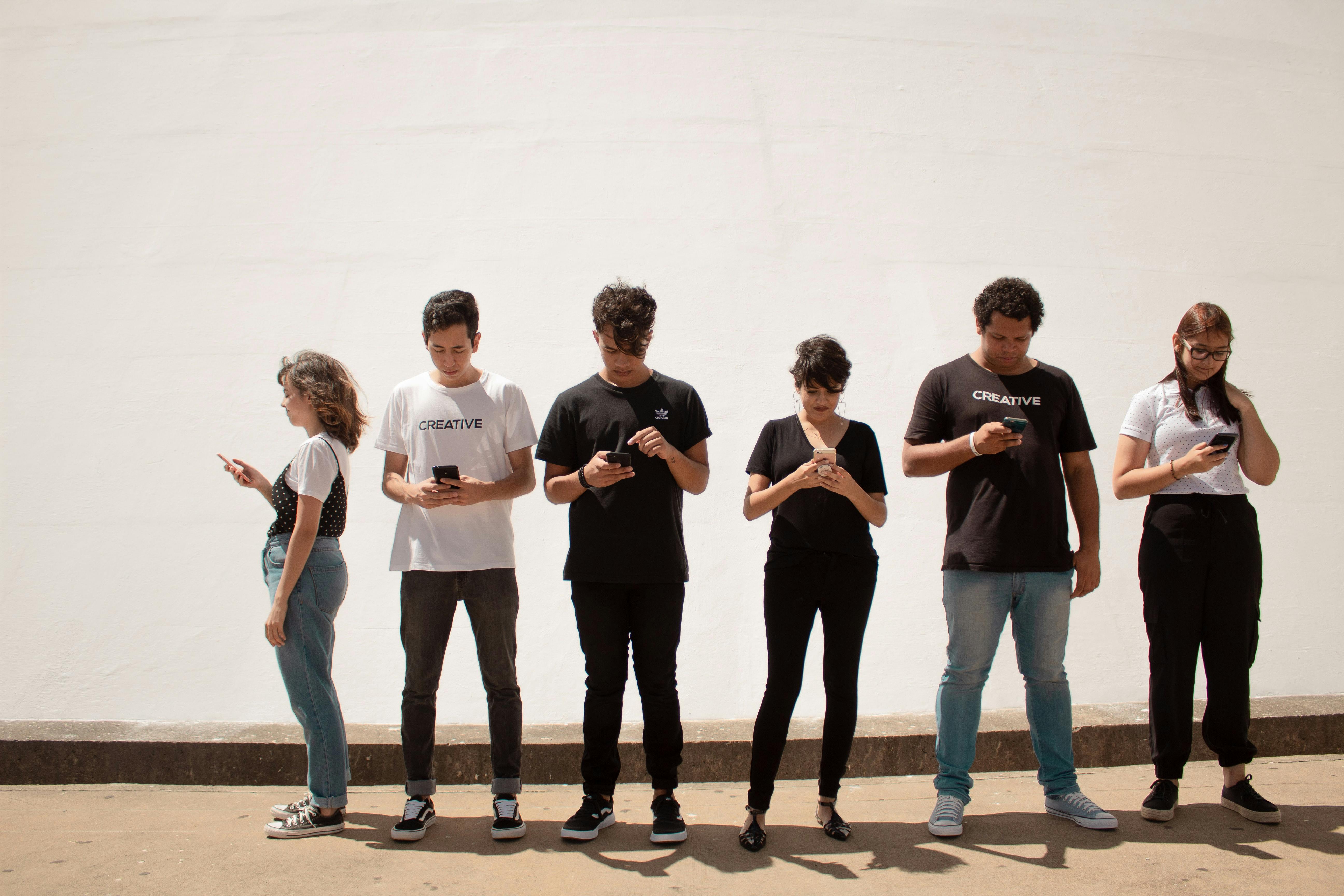 Six people are standing next to each other, all looking at their smartphone.