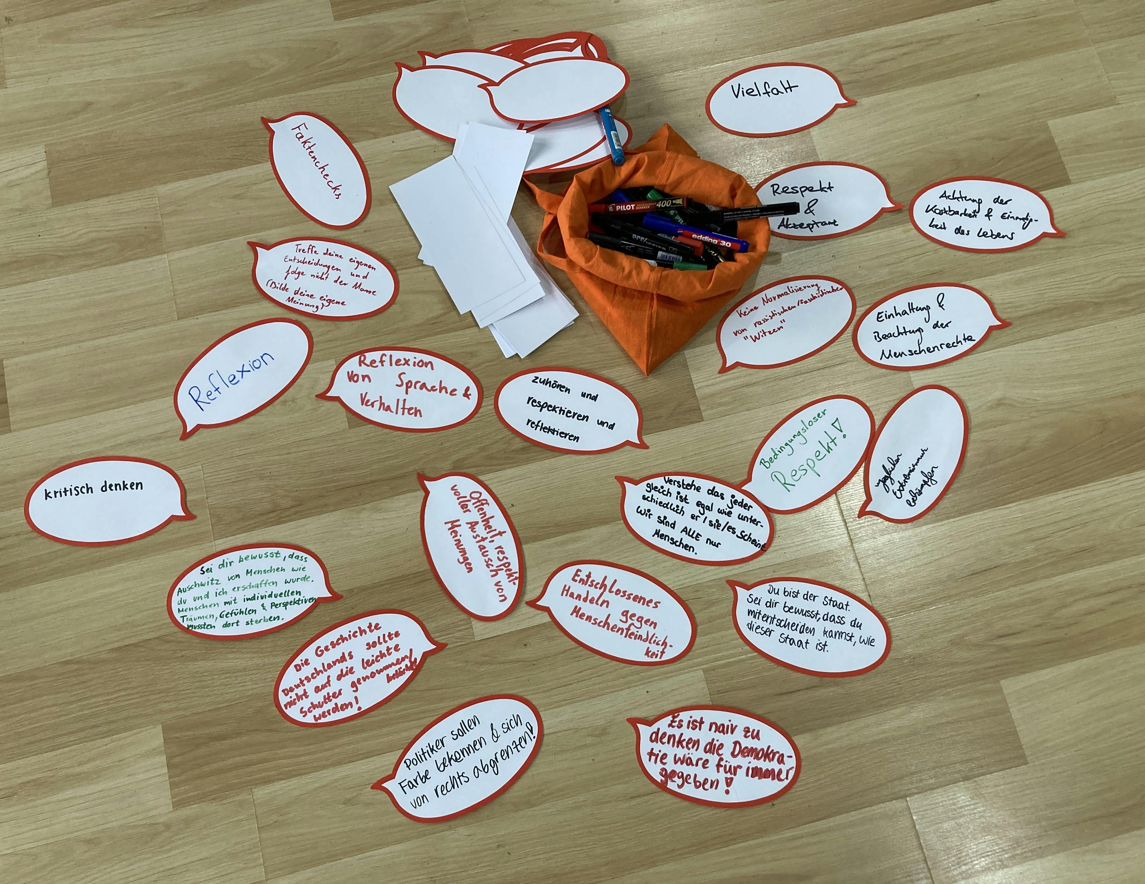 Auf dem Foto sieht man Papier-Sprechblasen, die bei der Gedenkstättenfahrt 2025 von Schüler:innen geschrieben wurden. Zwischen den Papierzetteln liegt ein orangener Stoffbeutel mit Markern darin. Im Vordergrund steht auf zwei der Zettel: "Politiker sollen Farbe bekennen & sich von rechts abgrenzen" oder "Es ist naiv zu glauben, die Demokratie sei für immer gegeben". 