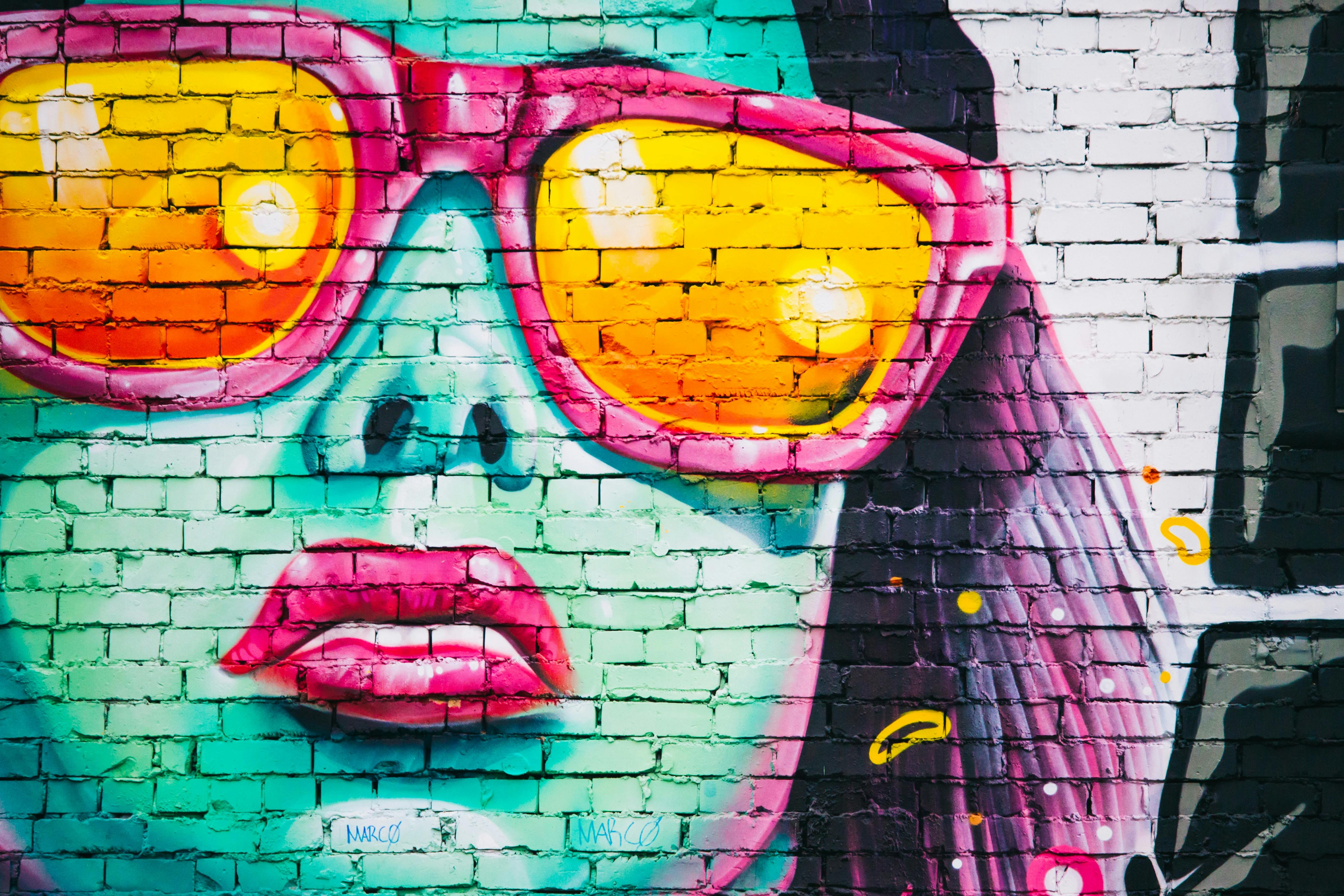 Es ist ein Graffiti mit einer weiblich positionierten Person zu sehen. SIe hat eine sehr coole Attitude und trägt eine Sonnenbrille. Dominierende Farben auf dem Graffiti sind Türkis, Pink und Gelb.