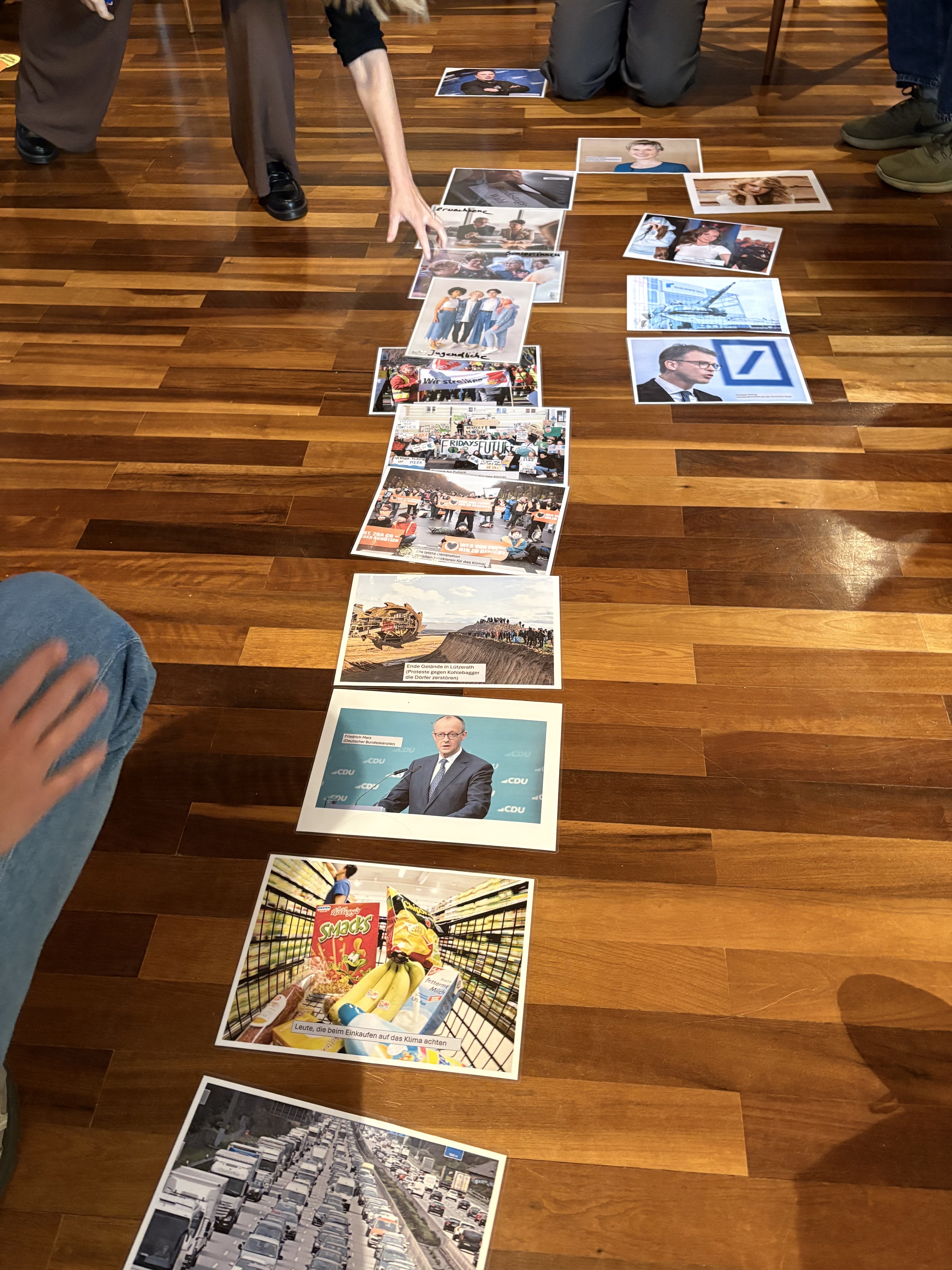 Auf einem braunen Parkettboden liegen Fotos von unter anderem einem Supermarkt, Friedrich Merz und Klimaaktivist:innen. Man sieht, dass darum Menschen sitzen oder knien, die diese in einer Linie anordnen. 