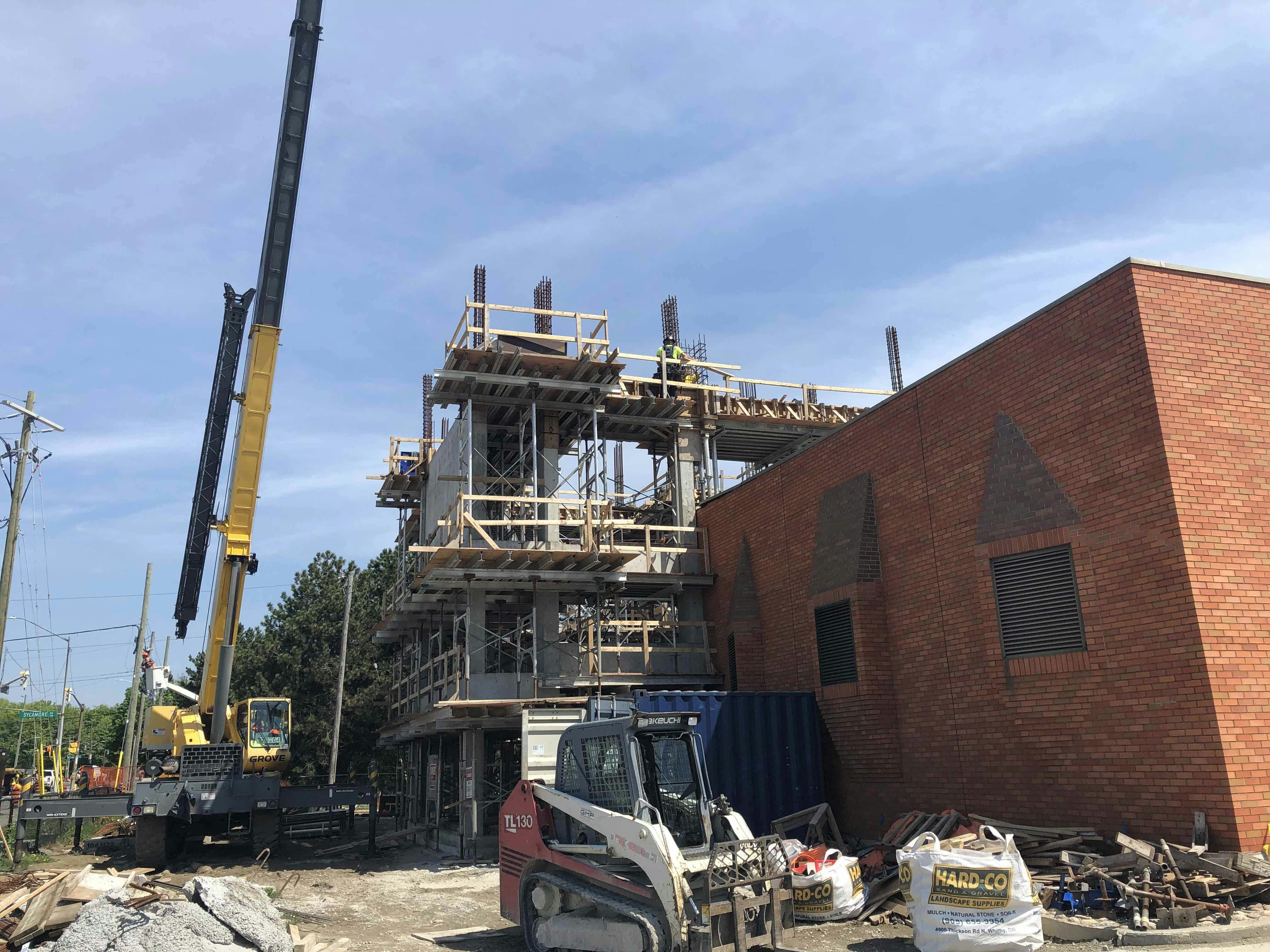 Construction site with a multi-story building under construction, a crane, and construction vehicles near a red brick building