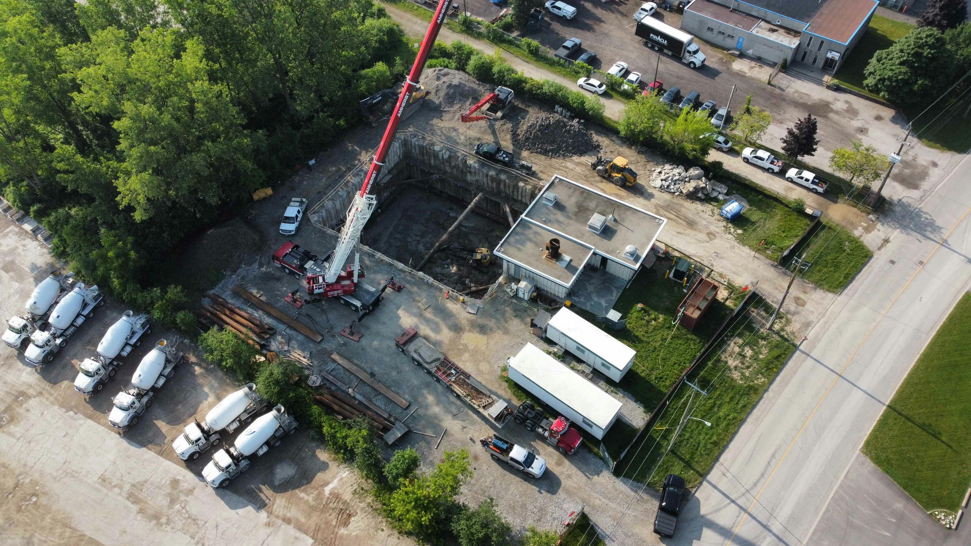 Aerial view of a construction site with a large excavated pit, a crane, construction vehicles, temporary buildings, and multiple cement trucks parked nearby