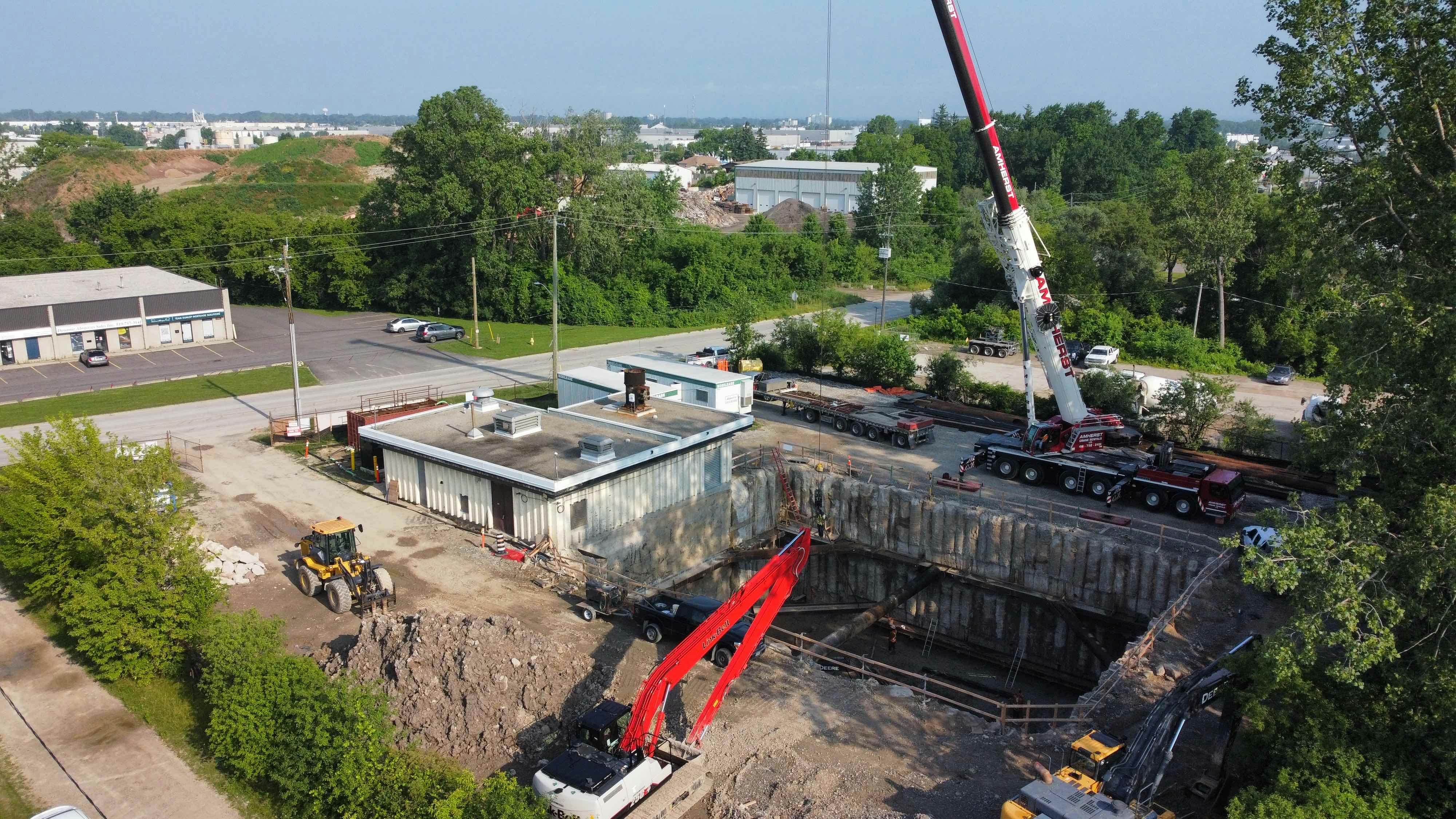 Ground-level view of a construction site with a large excavated pit, a crane, construction vehicles, and temporary buildings, surrounded by greenery and nearby industrial buildings