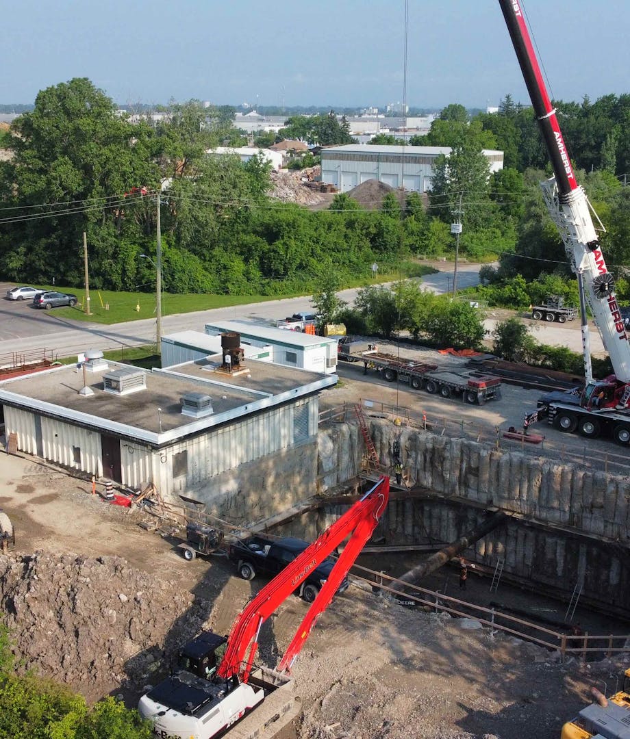 Ground-level view of a construction site with a large excavated pit, a crane, construction vehicles, and temporary buildings, surrounded by greenery and nearby industrial buildings