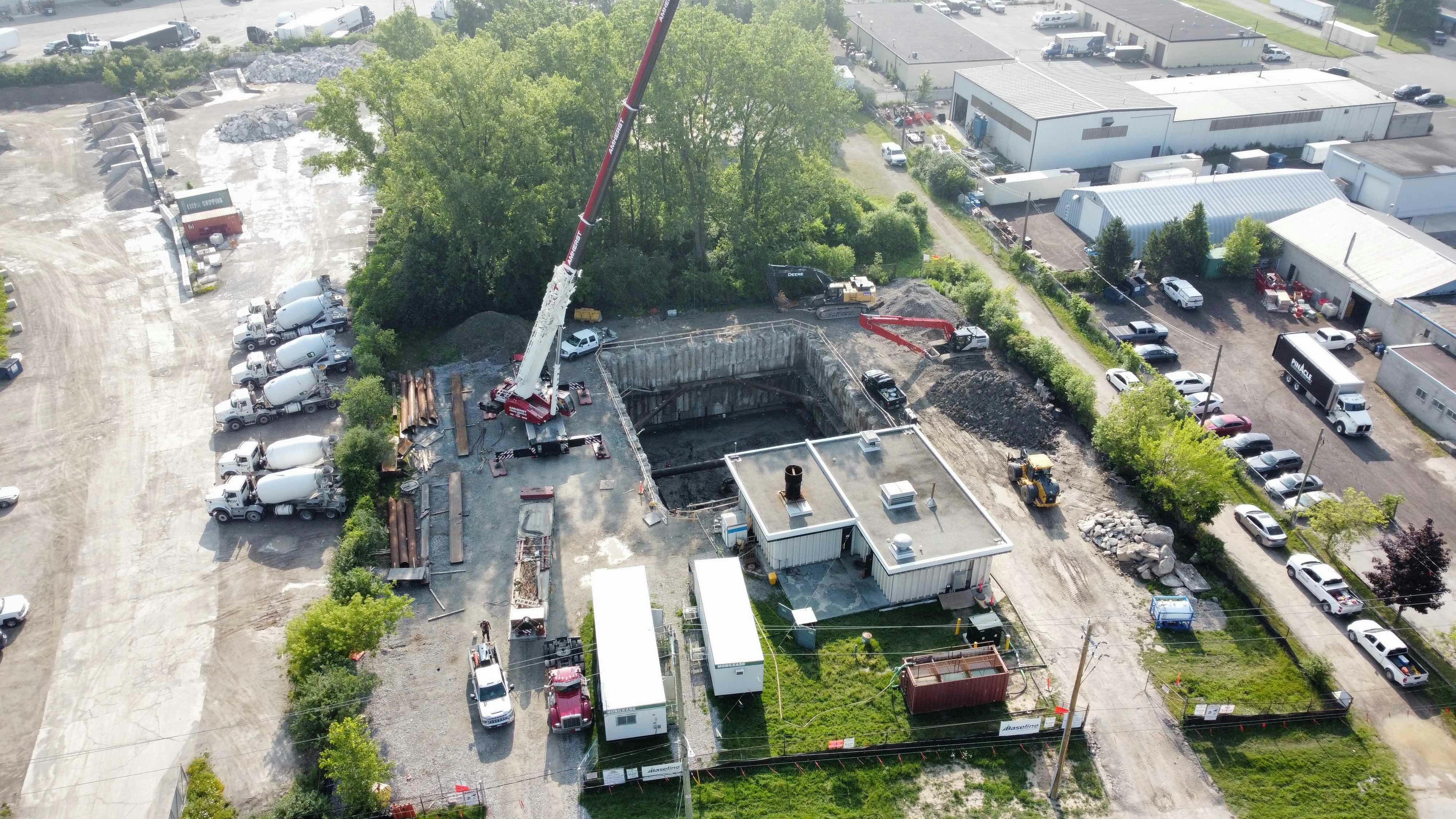 Aerial view of a construction site with a large excavated pit, a crane, construction vehicles, temporary buildings, and a row of cement trucks parked nearby