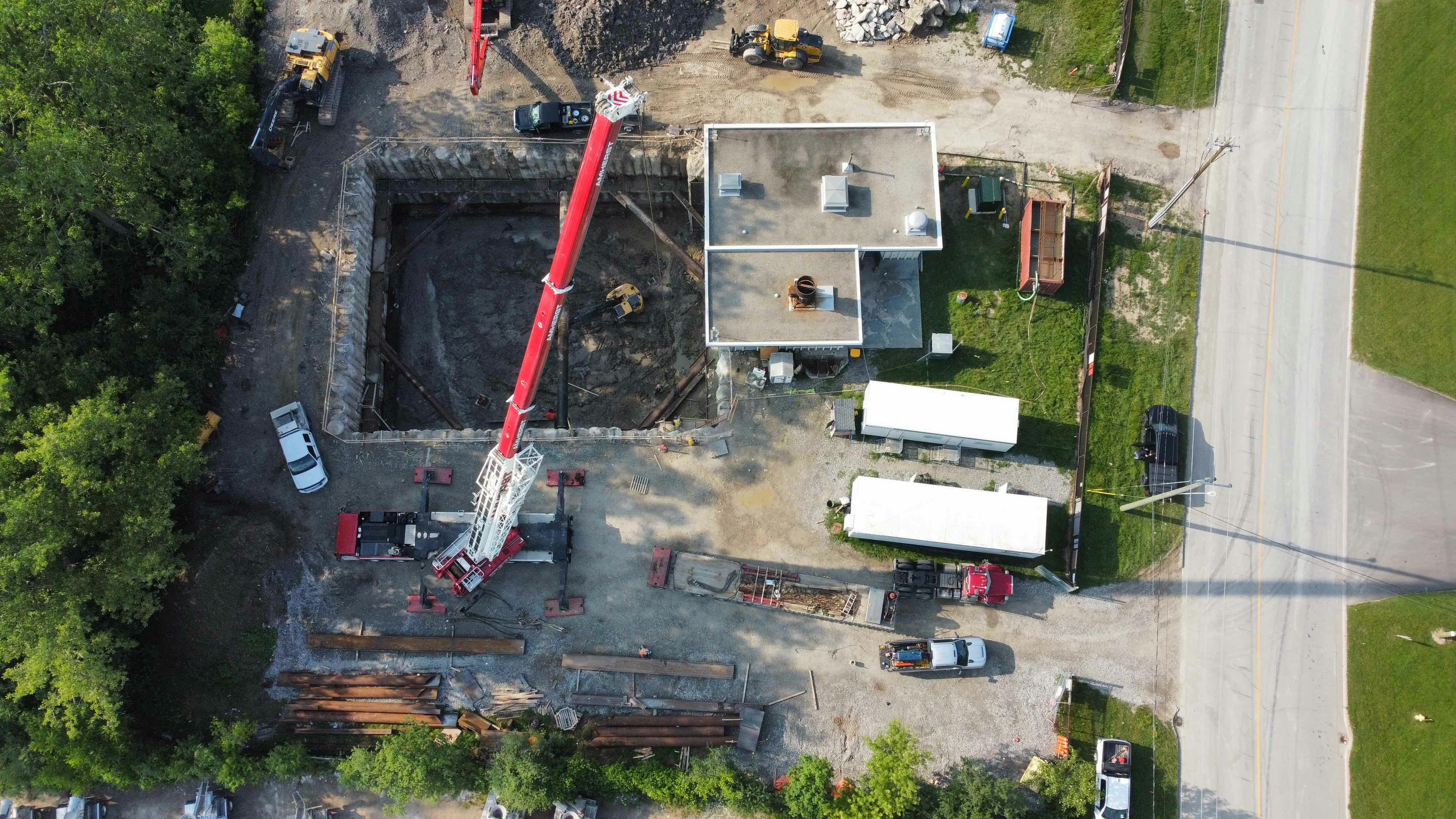 Aerial view of a construction site featuring a large excavated pit, a crane, construction vehicles, temporary buildings, and adjacent roads