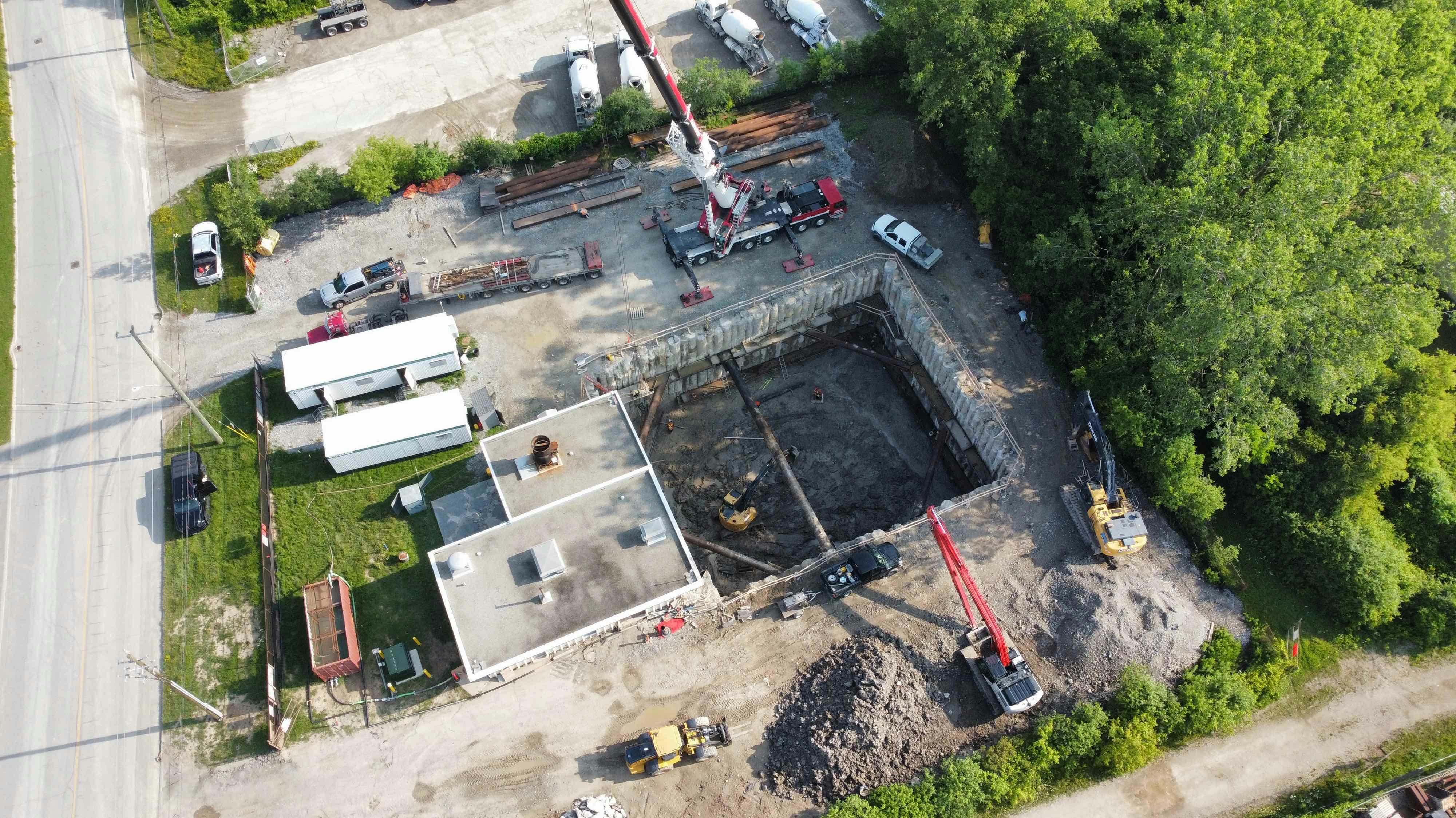 Aerial view of a construction site with a large excavated pit, a crane, construction vehicles, temporary structures, and surrounding greenery