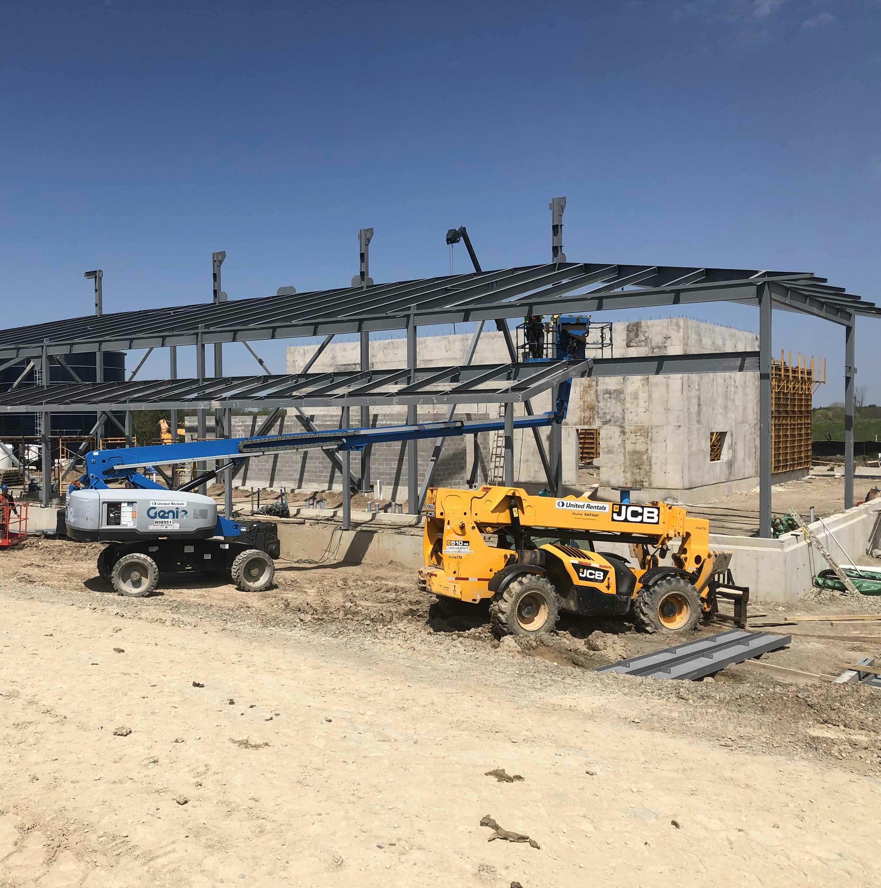 Construction site with a partially built structure, a yellow vehicle, a blue lift, and other construction equipment on a sandy ground