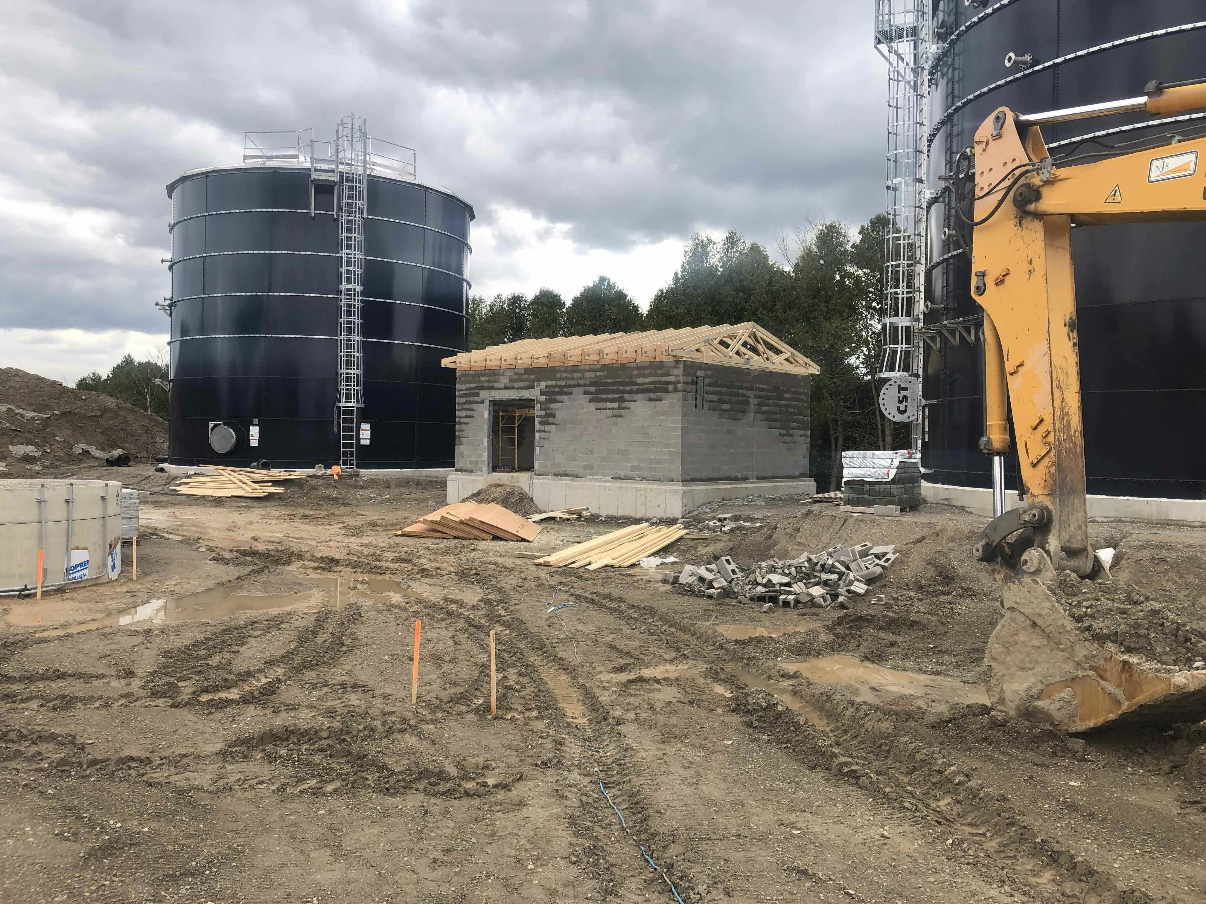 Construction site with a small unfinished building, large cylindrical tanks, and an excavator, set against a cloudy sky