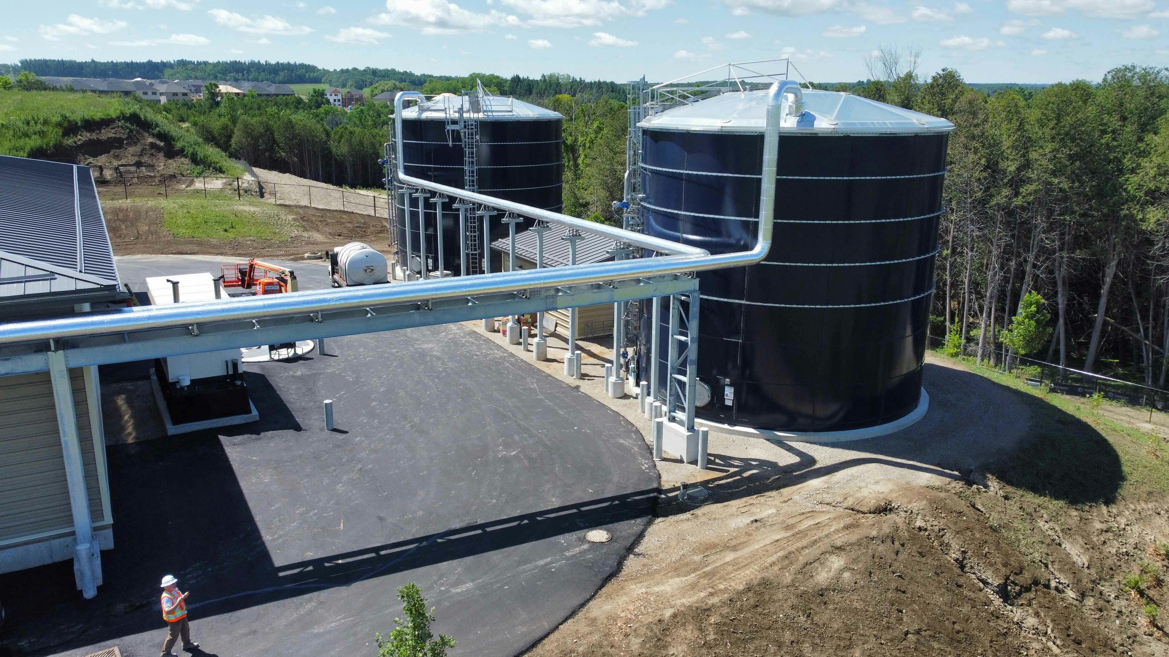 Ground-level view of an industrial site with two large cylindrical tanks connected by overhead pipes, a worker in a safety vest, and surrounding greenery