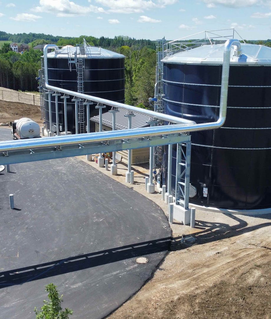 Ground-level view of an industrial site with two large cylindrical tanks connected by overhead pipes, a worker in a safety vest, and surrounding greenery