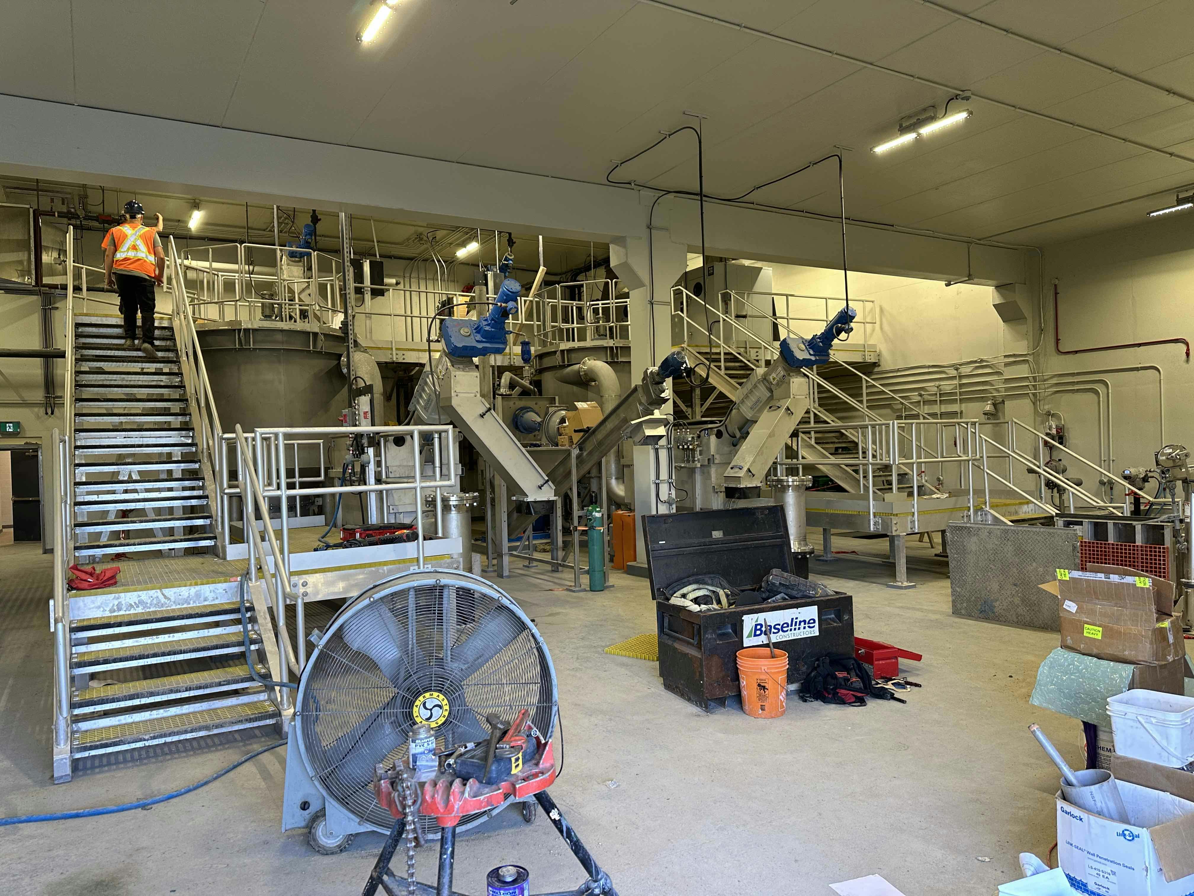 Interior of an industrial facility with metal staircases, machinery, a large fan, tools, and a worker in a safety vest