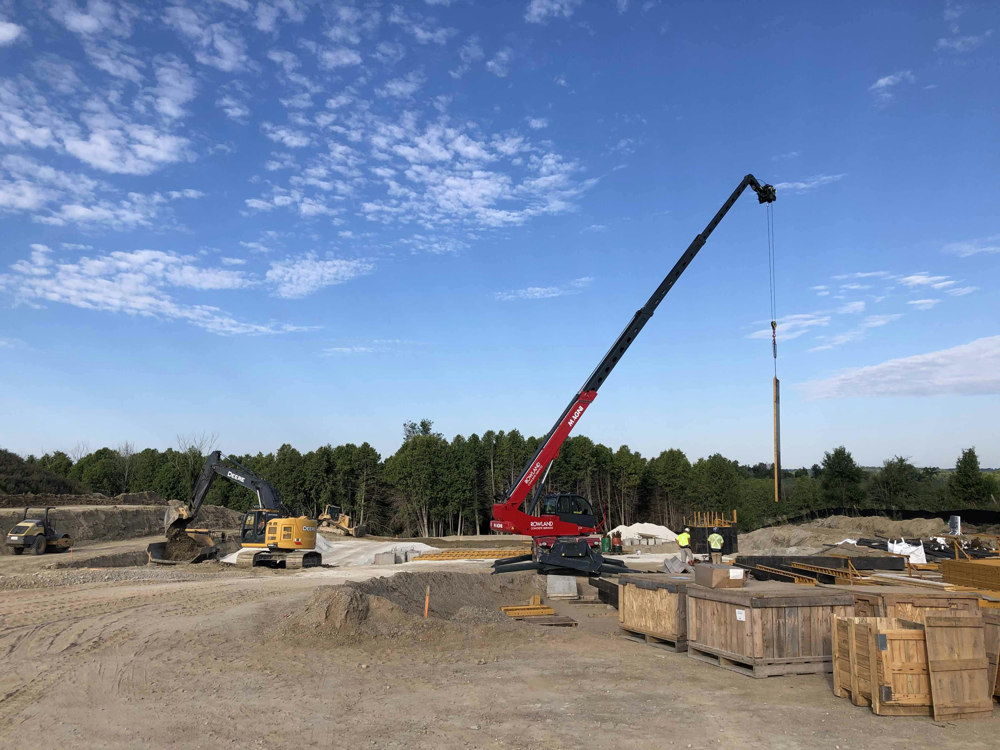 Construction site with a red crane lifting materials, an excavator, and wooden crates, set against a backdrop of trees and a blue sky