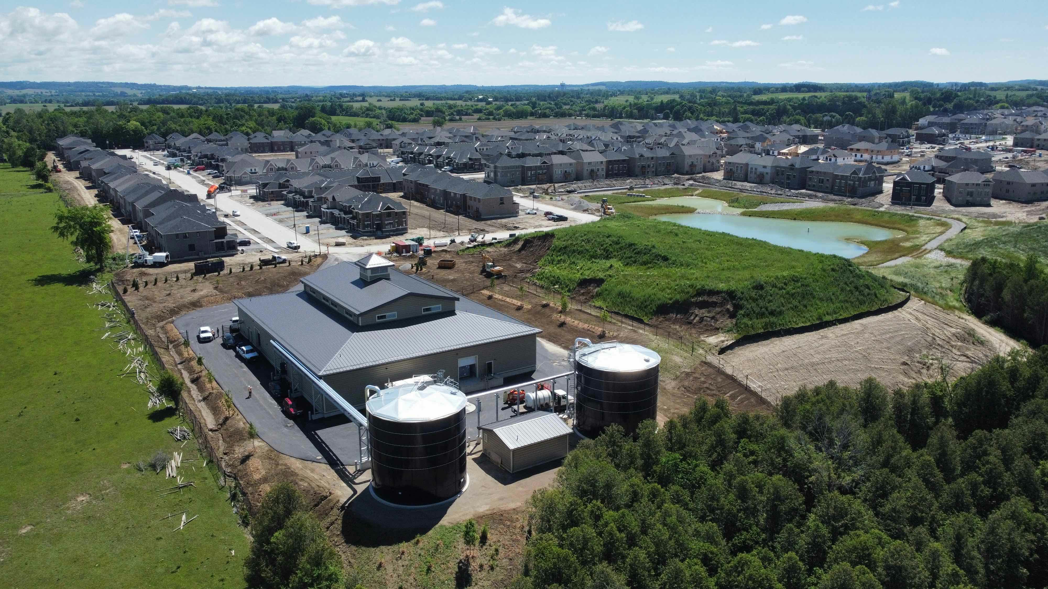 Aerial view of an industrial facility with two large cylindrical tanks, surrounded by greenery, residential buildings, and a small pond