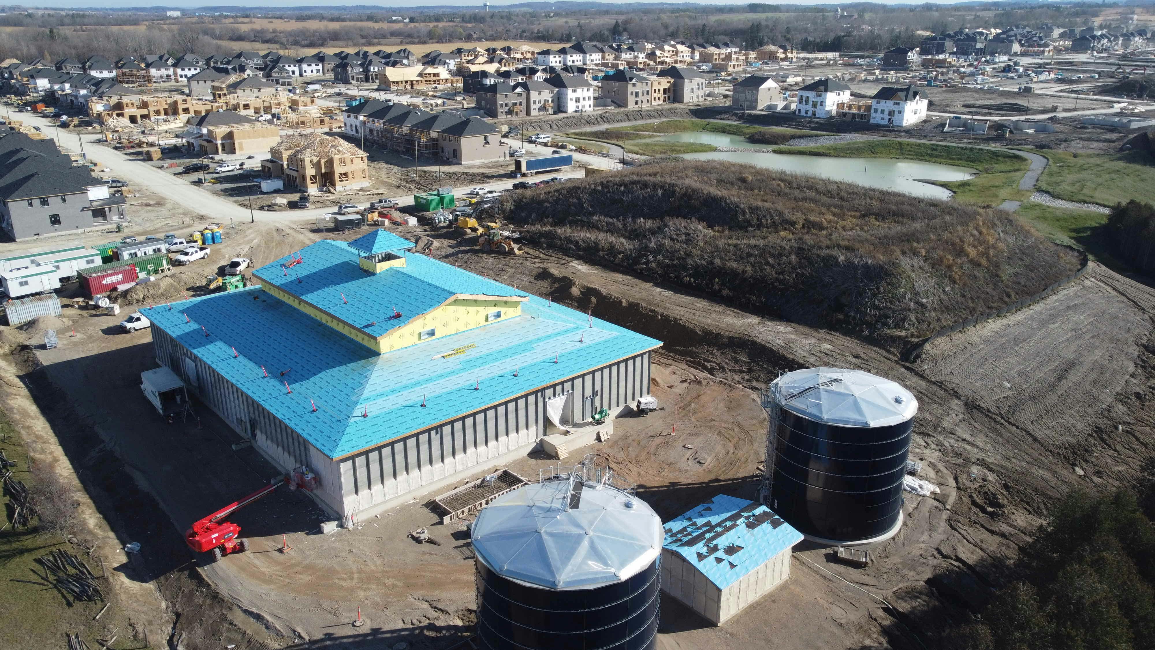 Aerial view of a construction site with a large building with a blue roof, two large cylindrical structures, surrounding dirt paths, and nearby residential buildings