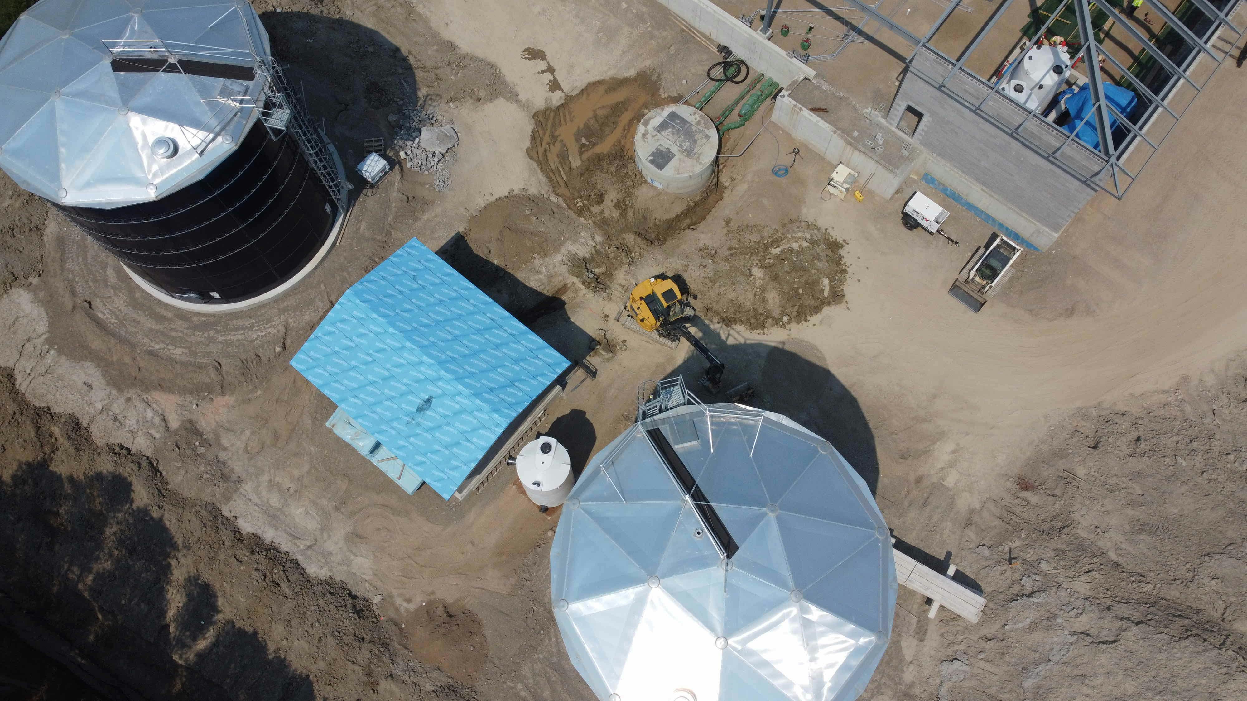 Aerial view of an industrial site with two large domed structures, a blue-roofed building, a construction vehicle, and surrounding dirt paths