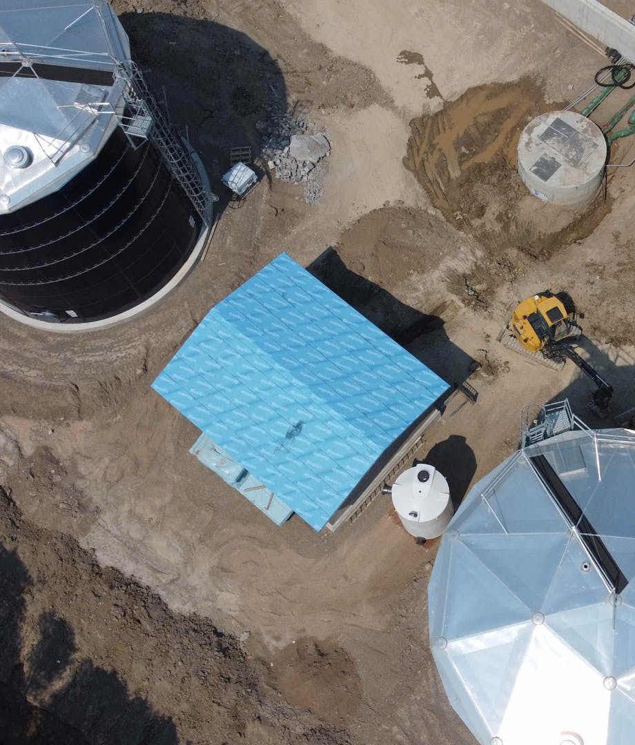 Aerial view of an industrial site with two large domed structures, a blue-roofed building, a construction vehicle, and surrounding dirt paths