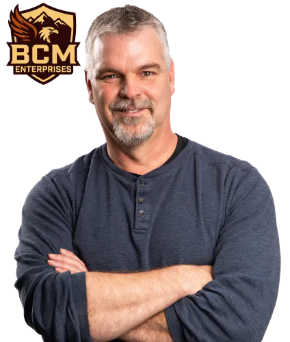 Shield badge showing a mountain and pine tree landscape with the text “BCM Enterprises” in gold and brown beside a photo of Bernie McClean with his arms crossed