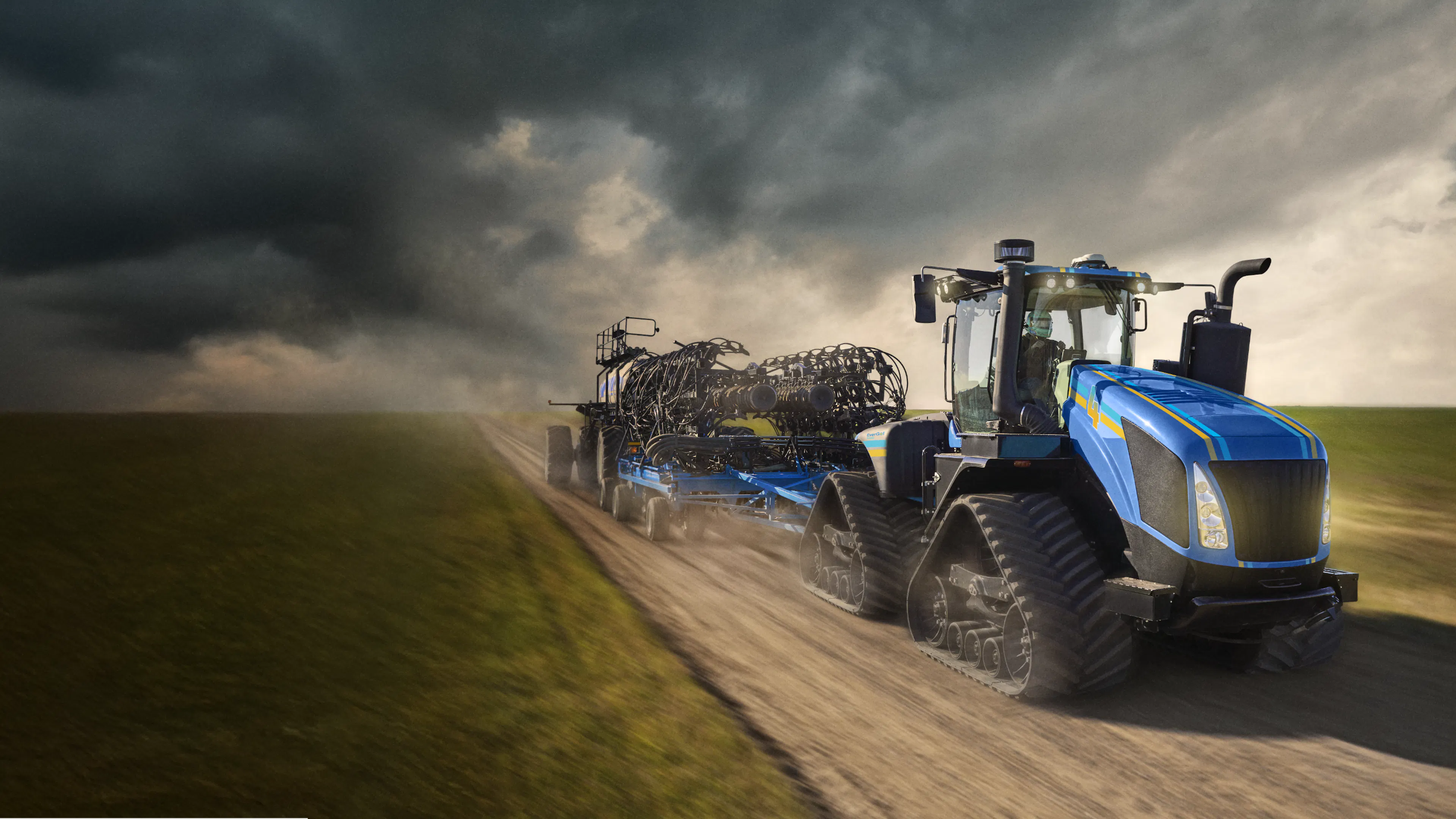 A large blue tractor pulls a piece of seeding equipment down a dirt road, kicking up dust as it moves through an open rural landscape under a cloudy sky. 