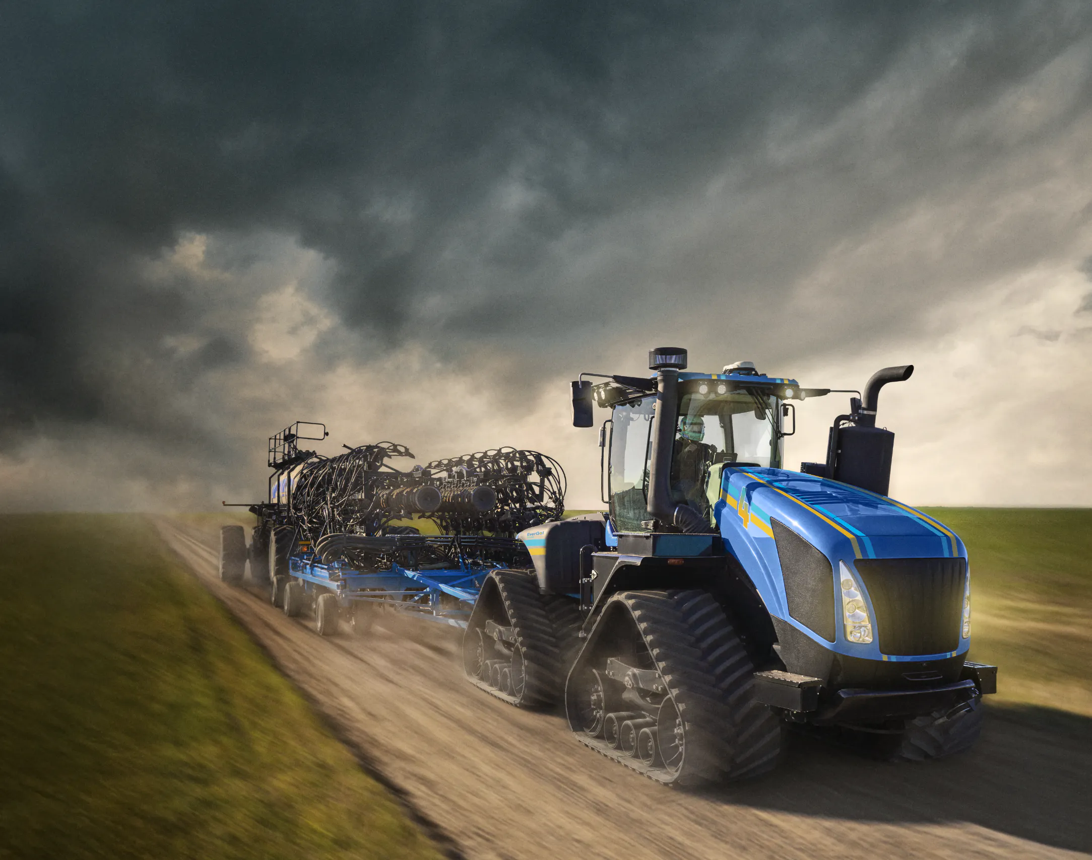 A large blue tractor pulls a piece of seeding equipment down a dirt road, kicking up dust as it moves through an open rural landscape under a cloudy sky.