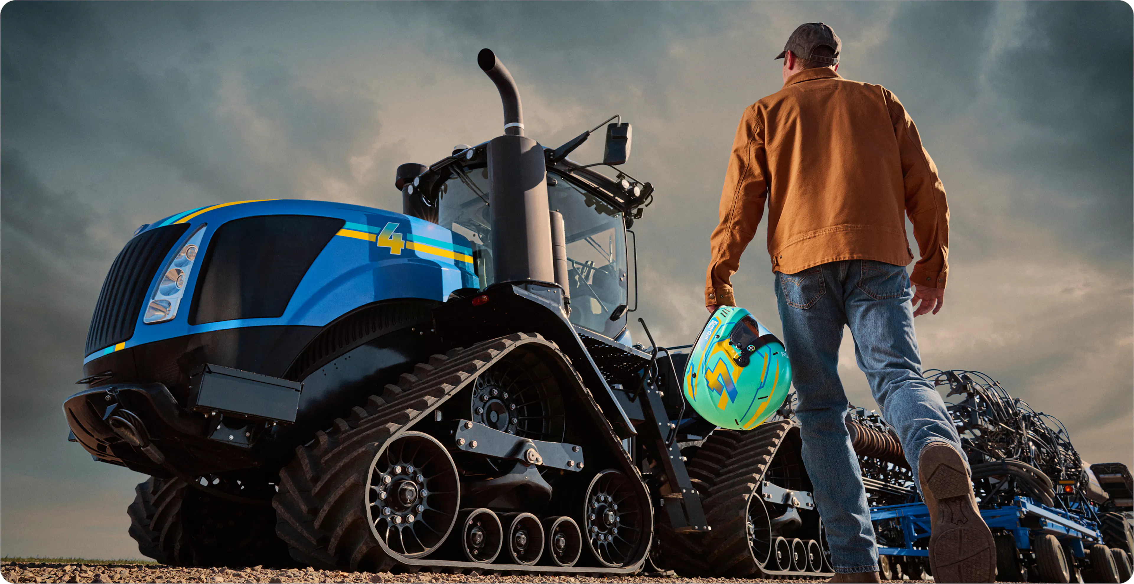 A person wearing a brown jacket and jeans walks toward a large blue tracked tractor, holding a brightly colored helmet in one hand.