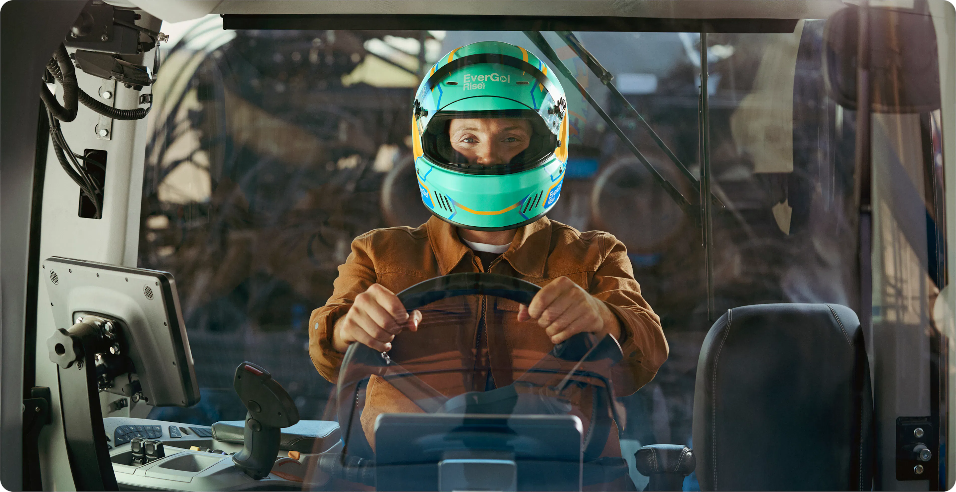 A person wearing a brightly colored racing helmet sits inside the cab of a large agricultural machine, gripping the steering wheel with both hands and looking forward through the front window. 