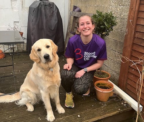 A person in a purple Blood Cancer UK shirt kneels on a wooden deck beside a light-coloured dog, with potted plants and outdoor furniture in the background.