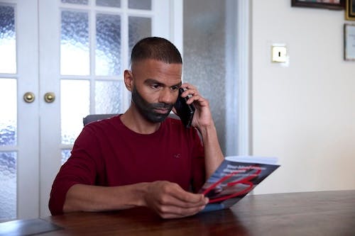 A person in a red long-sleeve top sits at a table, speaking on the phone while holding an informational booklet.