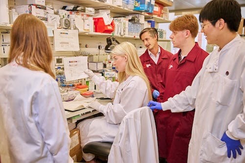 A group of researchers in lab coats gather around a colleague who is seated at a lab bench reviewing notes and equipment.