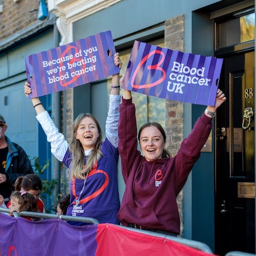 Two people standing behind a barrier holding signs that read ‘Because of you we’re beating blood cancer’ and ‘Blood Cancer UK’ at an outdoor event.