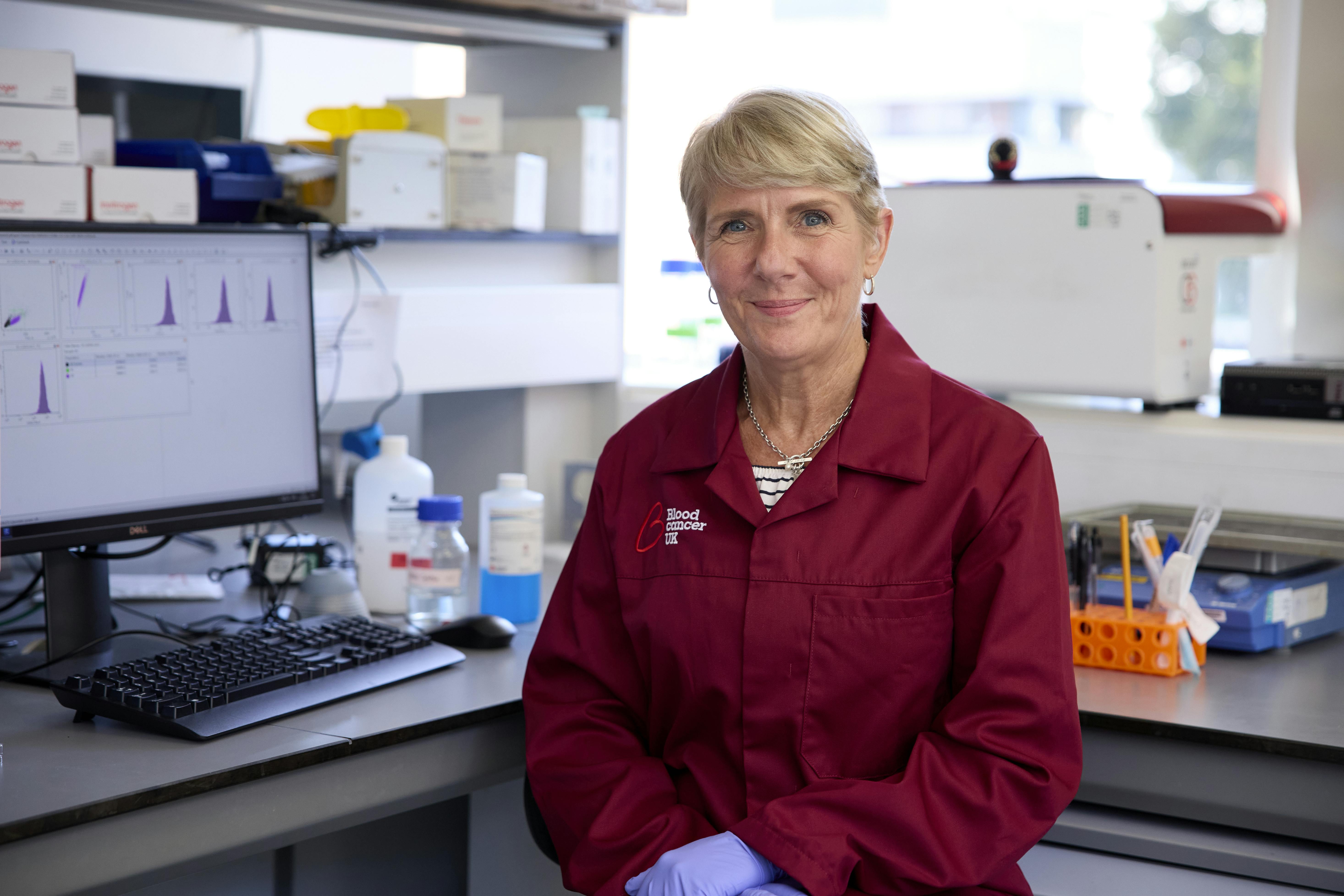 Photo of a researcher wearing a burgundy lab coat, sitting in a lab.