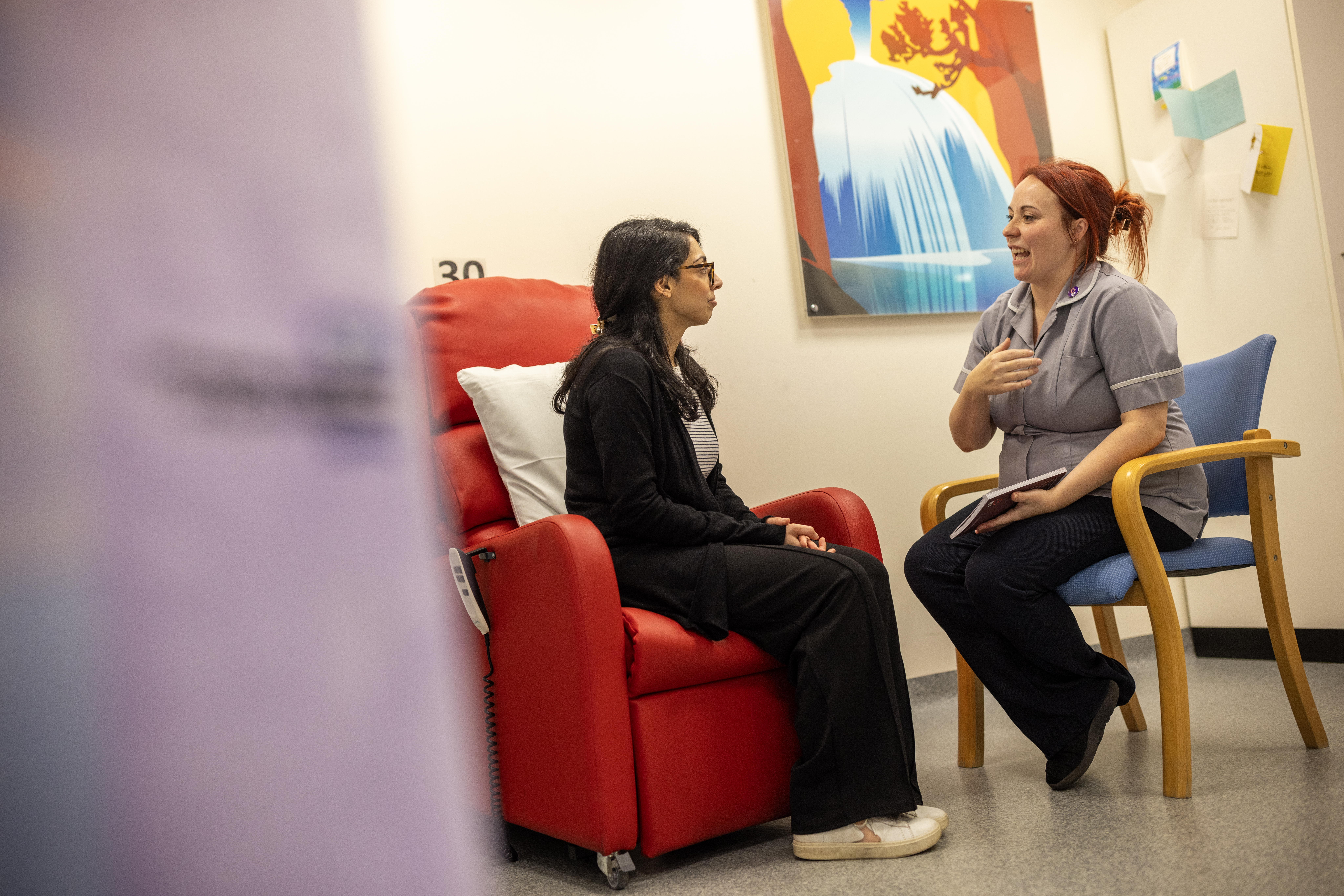 Clinical trial nurse and patient seated in a waiting room, chatting.