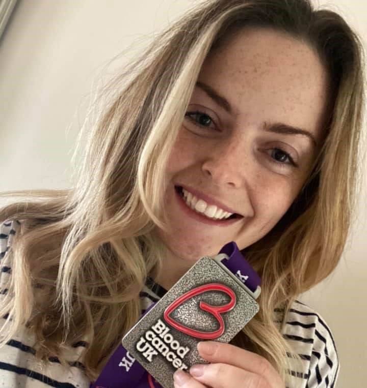 A woman with blonde hair holding a Blood Cancer UK medal up and smiling