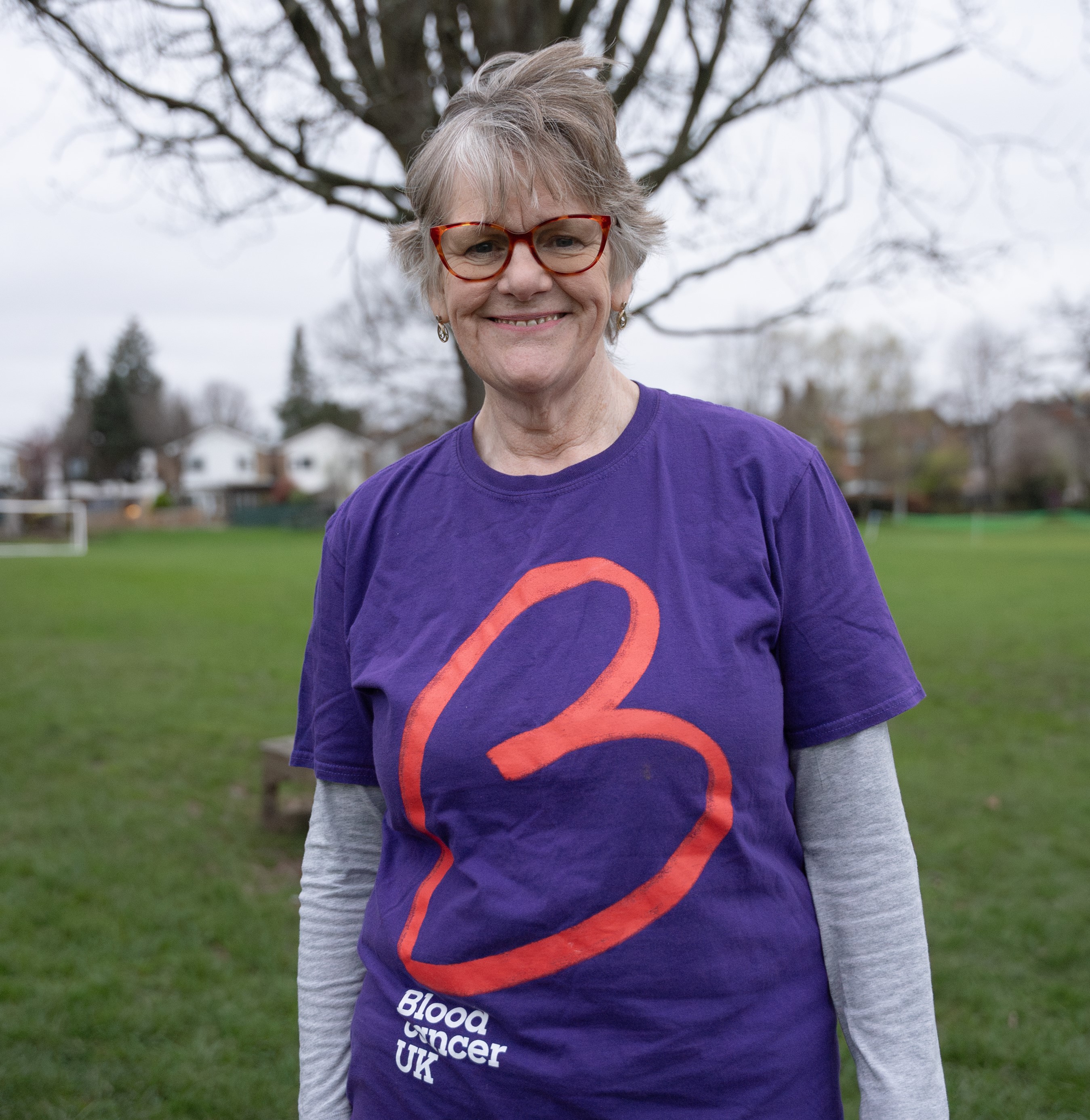 A woman standing outside in a field in front of a tree with no leaves, with grey hair and red rimmed glasses smiling wearing a purple t-shirt with a red big ‘B’ logo on the front