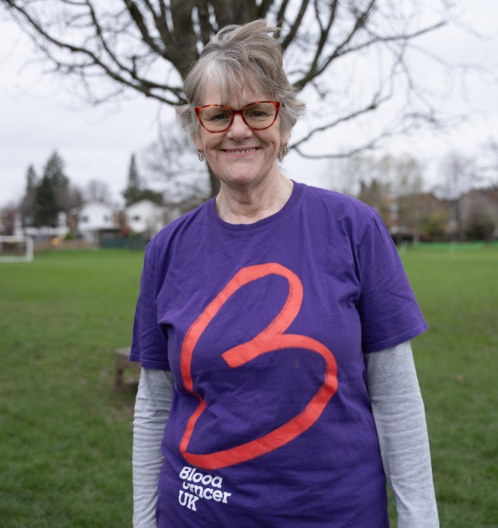 A woman standing outside in a field in front of a tree with no leaves, with grey hair and red rimmed glasses smiling wearing a purple t-shirt with a red big ‘B’ logo on the front