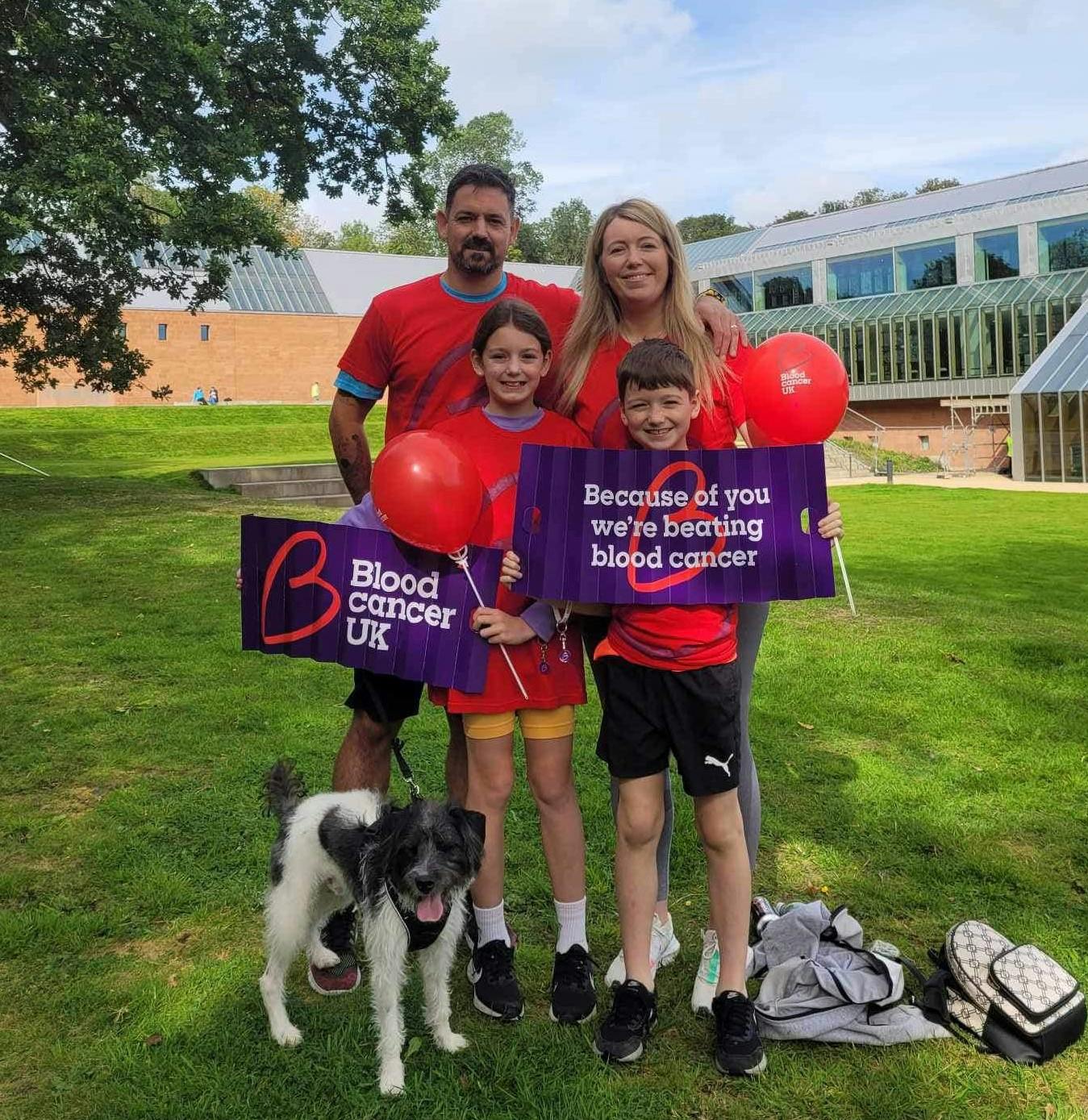 A mum, dad, son and daughter standing outside a greenhouse on the grass with their dog holding balloons and signs that say ‘Blood Cancer UK’ and ‘Because of you we’re beating blood cancer’