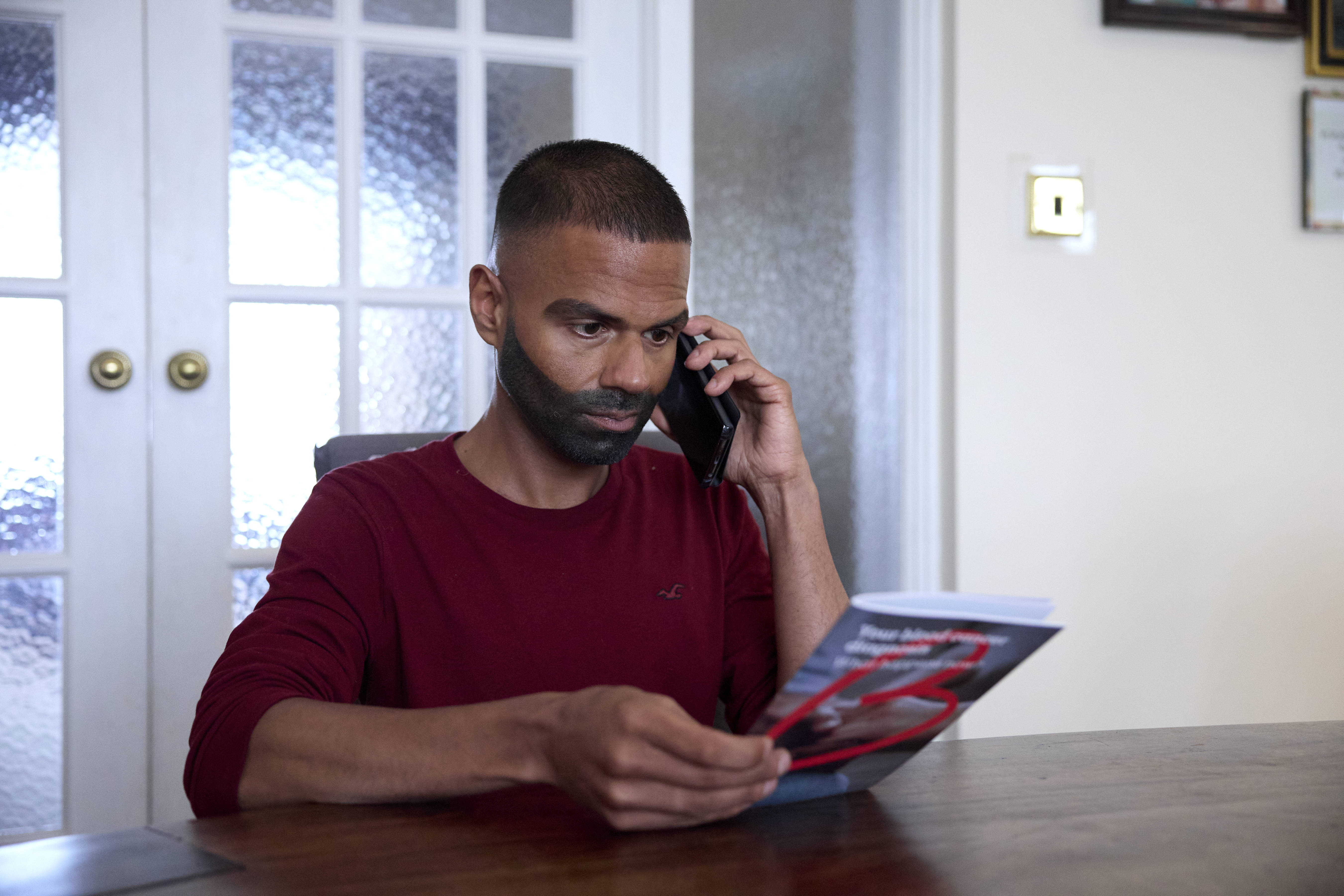 A man sitting at the table holding a blood cancer information booklet and holding his phone up with a concentrated look on his face.