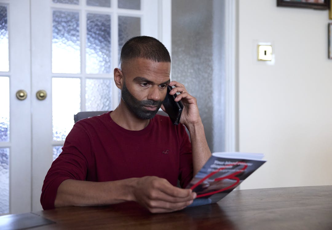A man sitting at the table holding a blood cancer information booklet and holding his phone up with a concentrated look on his face.