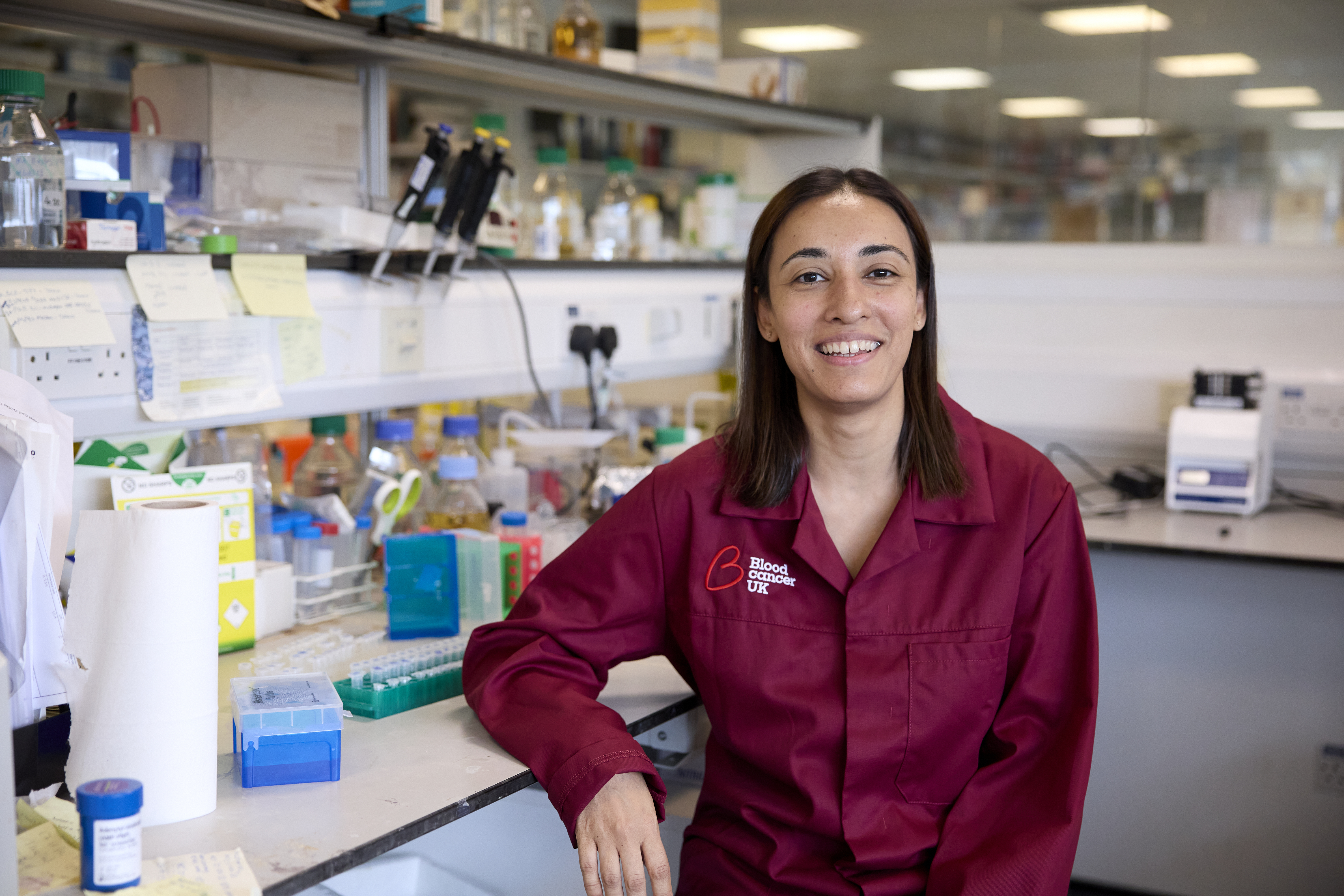 A researcher sitting in a lab, smiling with equipment behind her and wearing a burgundy lab coat