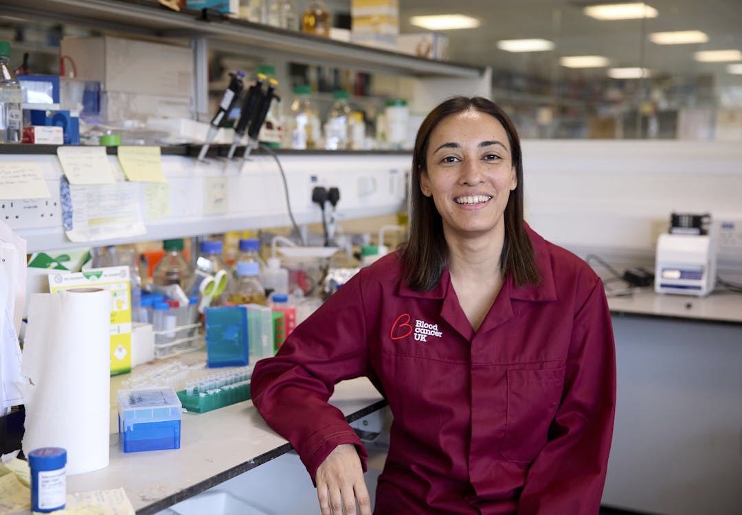 A researcher sitting in a lab, smiling with equipment behind her and wearing a burgundy lab coat