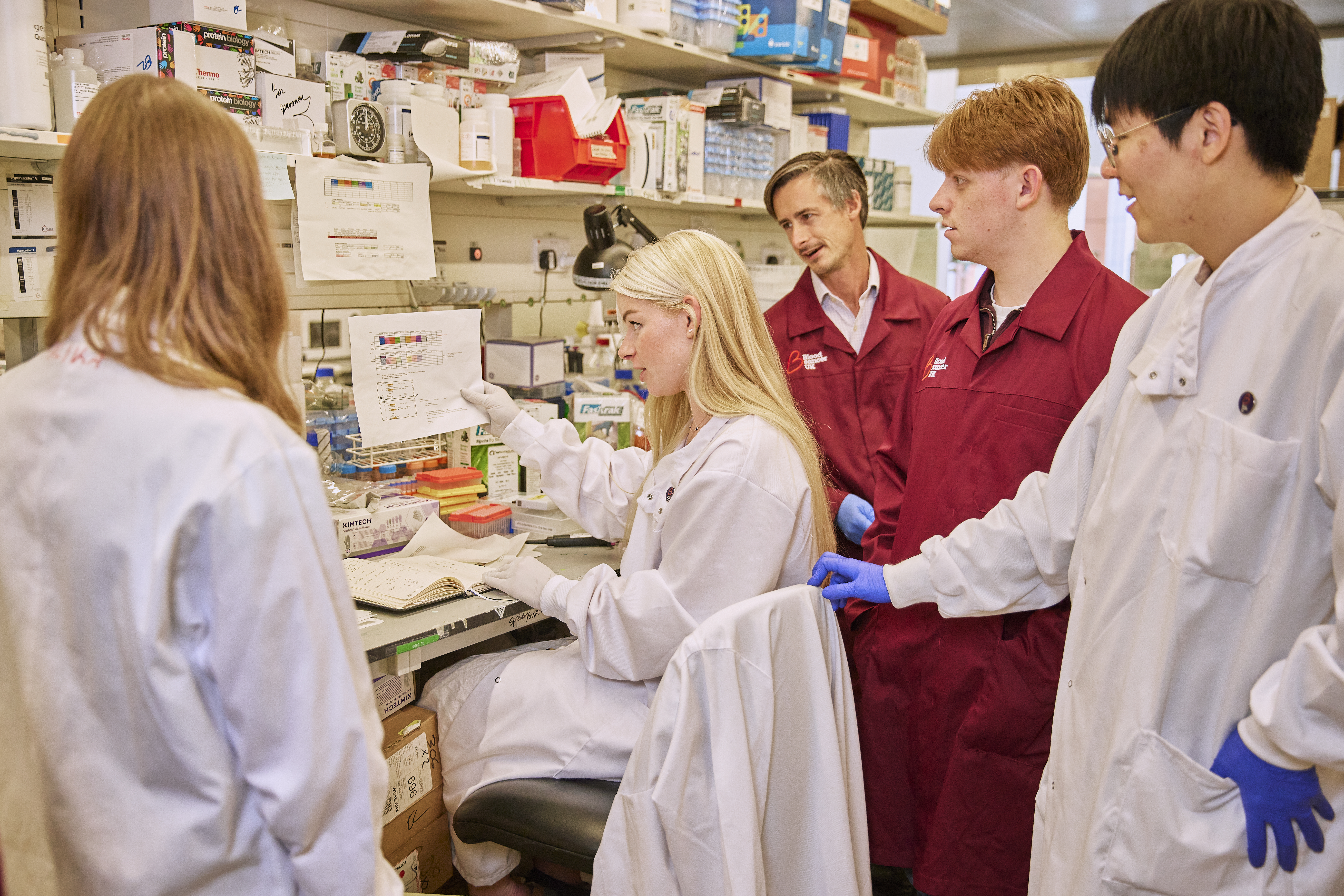 Four researchers standing around another researcher in the lab who is sat down explaining a diagram on a piece of paper to them