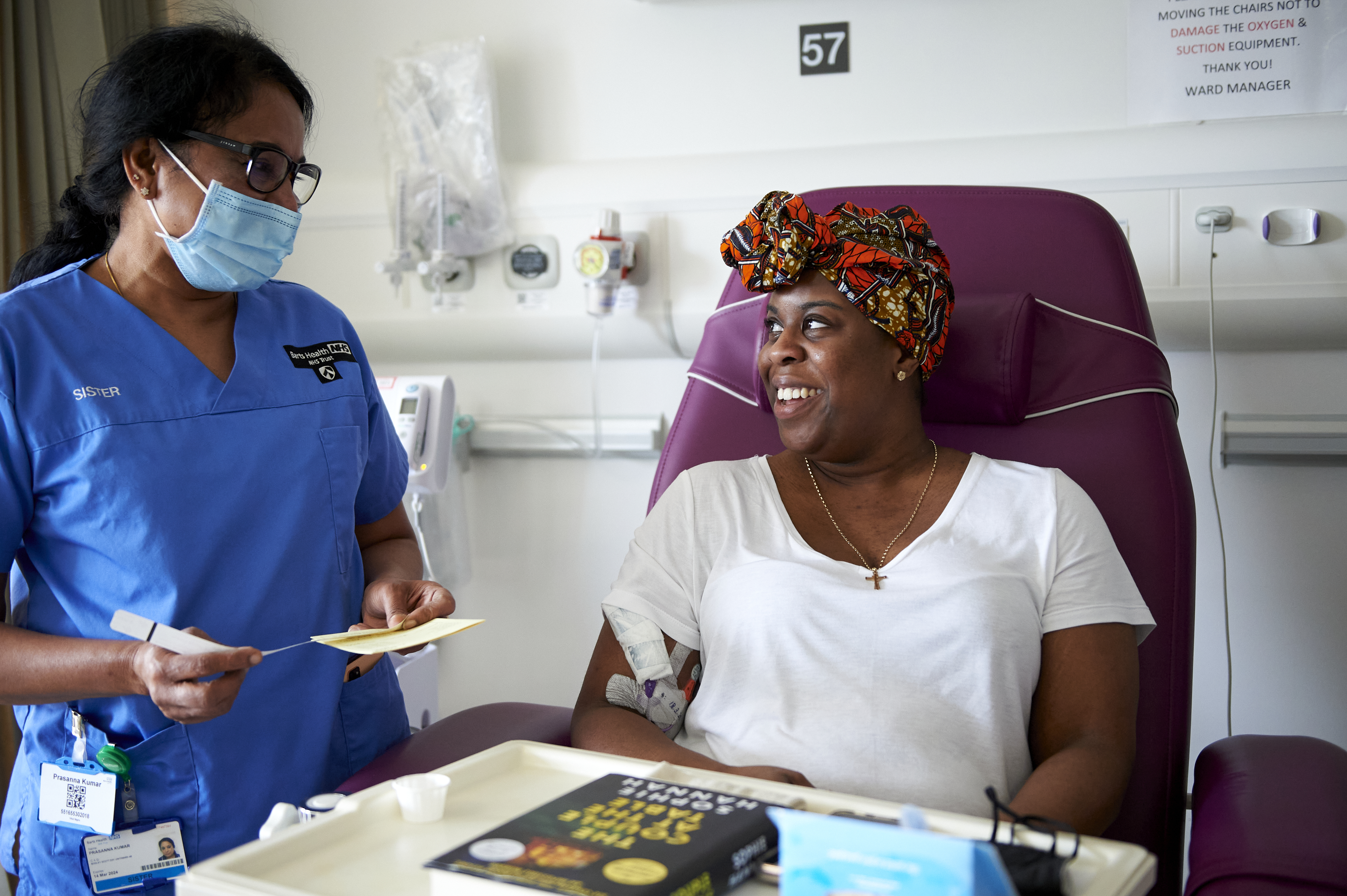 A clinical nurse specialist standing next to a hospital bed wearing her blue uniform and a blue face mask talking to a patient who’s sat in a purple chair with a drip in their arm